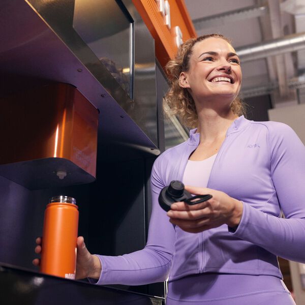 Femme en tenue de sport violette souriant tout en tenant une gourde orange Basic-Fit &agrave; c&ocirc;t&eacute; d&rsquo;une station d&rsquo;eau sportive Yanga dans une salle de sport.