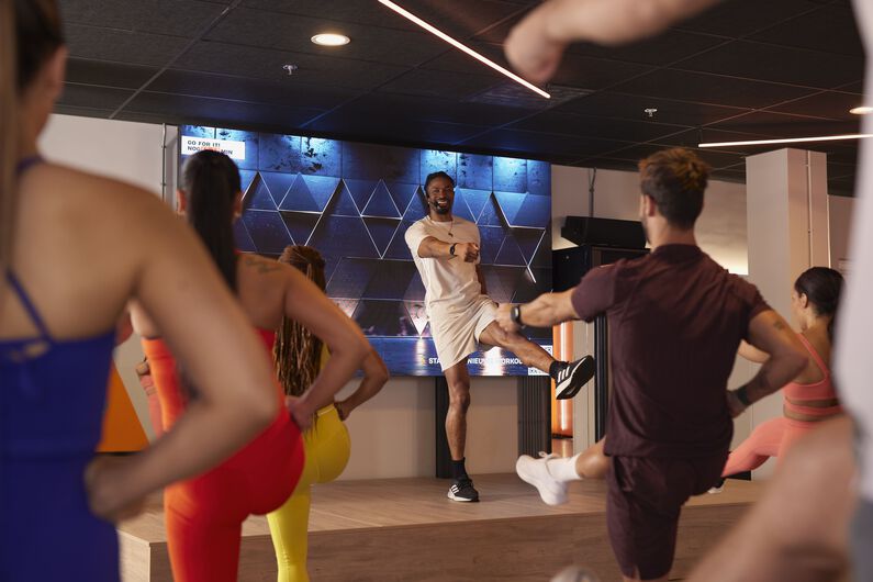 Cours collectif de fitness avec un instructeur sur une sc&egrave;ne montrant un mouvement, tandis que les participants s&rsquo;entra&icirc;nent au sol.