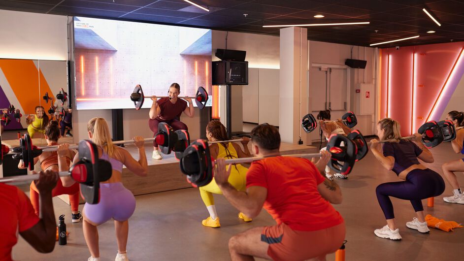 un groupe de personnes effectue des squats avec des barres pendant qu&rsquo;un instructeur montre le mouvement sur une sc&egrave;ne sur&eacute;lev&eacute;e dans une salle de sport