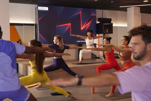 Group of people taking part in a yoga style group class in a gym studio, holding a wide standing pose with arms extended while an instructor leads the class from a platform.