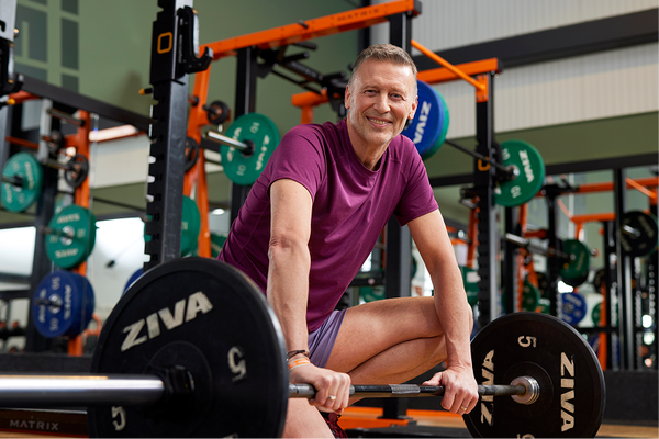 A middle-aged man smiles while preparing for a barbell lift in a gym, kneeling on one knee in front of a squat rack with weight plates and strength equipment in the background.