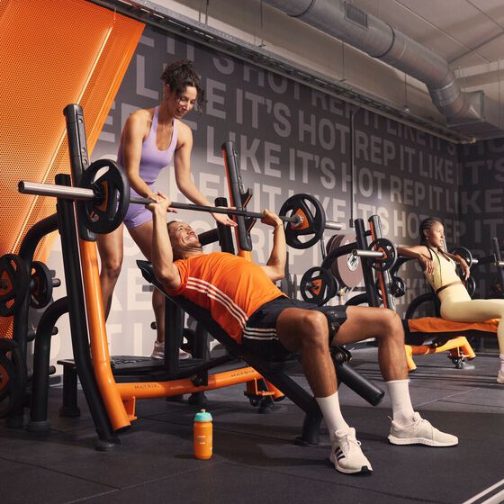 Un membre r&eacute;alise un d&eacute;velopp&eacute; inclin&eacute; avec l'aide d'un coach chez Basic-Fit, tandis que d'autres s'entra&icirc;nent sur des machines de musculation en arri&egrave;re-plan.