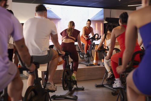 Un groupe de personnes p&eacute;dale sur des v&eacute;los stationnaires pendant qu&rsquo;une instructrice roule sur un v&eacute;lo sur une plateforme sur&eacute;lev&eacute;e &agrave; l&rsquo;avant de la salle.