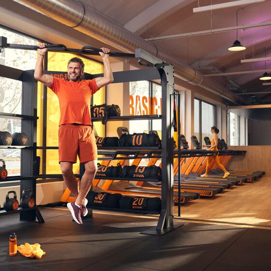 Un membre r&eacute;alise une traction sur une structure d'entra&icirc;nement fonctionnel chez Basic-Fit, tandis qu'un autre marche sur un tapis de course.