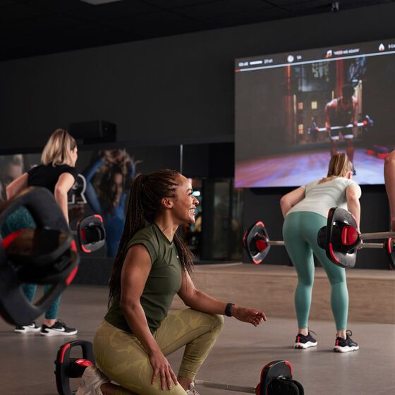 Un groupe de membres suit un entra&icirc;nement de musculation virtuel sur l'&eacute;cran GXR chez Basic-Fit et r&eacute;alise des exercices avec une barre.