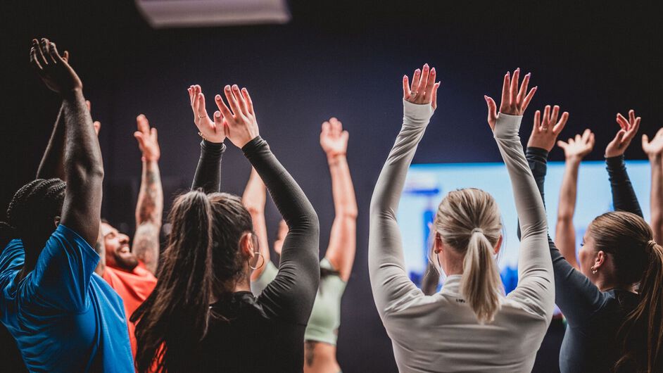 Un groupe de membres suit ensemble un entra&icirc;nement vid&eacute;o chez Basic-Fit et l&egrave;ve les bras de mani&egrave;re synchronis&eacute;e en faisant face &agrave; l&rsquo;&eacute;cran dans le studio.