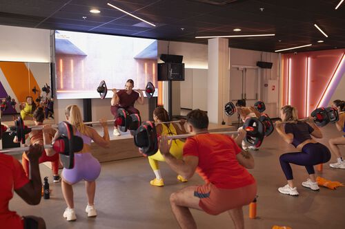 Un groupe de personnes effectue des squats avec des barres pendant qu&rsquo;une instructrice montre le mouvement sur une sc&egrave;ne sur&eacute;lev&eacute;e dans une salle de sport.