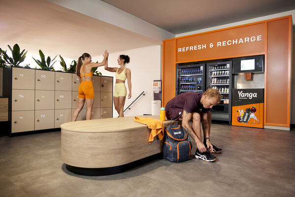 Des membres se pr&eacute;parent pour leur s&eacute;ance dans l&rsquo;espace vestiaire d&rsquo;un club Basic-Fit tandis que deux sportifs se saluent.