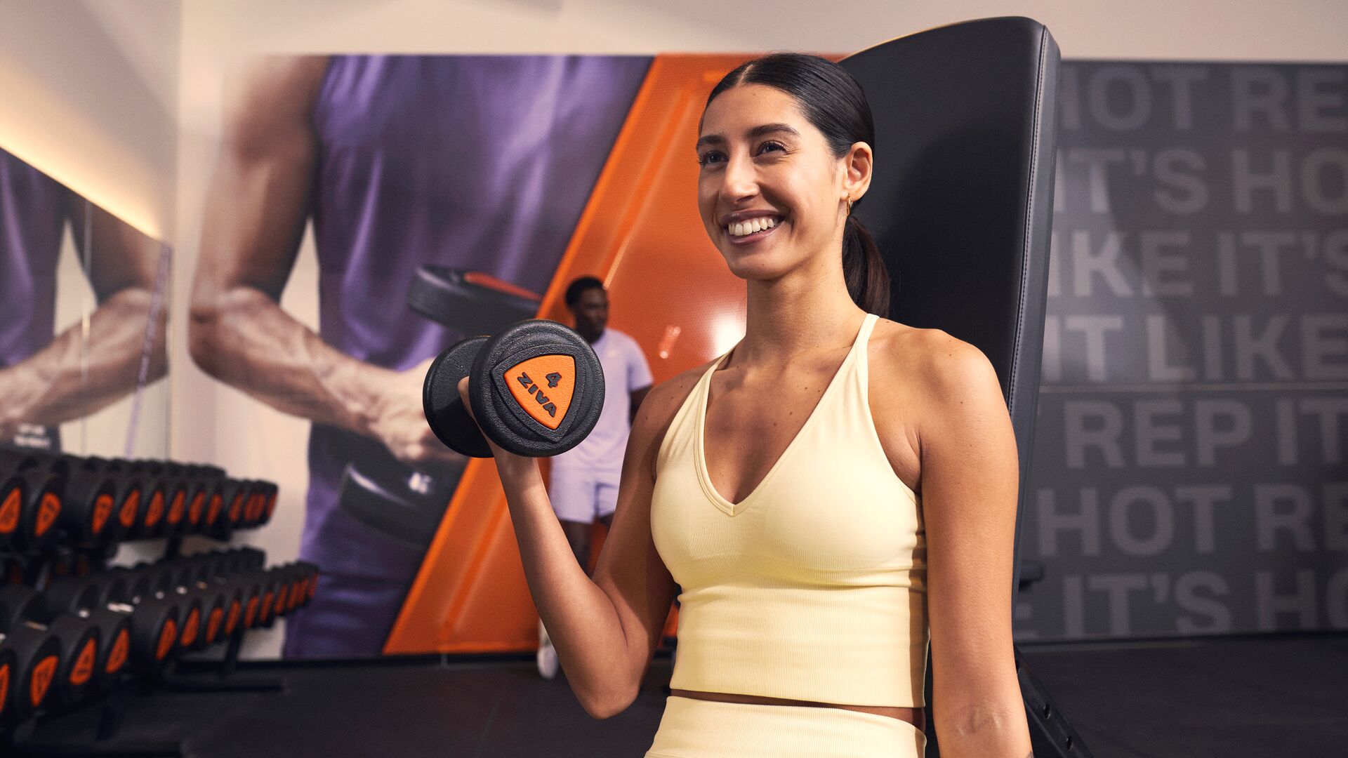 Personne effectuant un curl biceps avec un haltère dans une salle de sport, souriante, avec un rack d’haltères et des machines de musculation en arrière-plan.