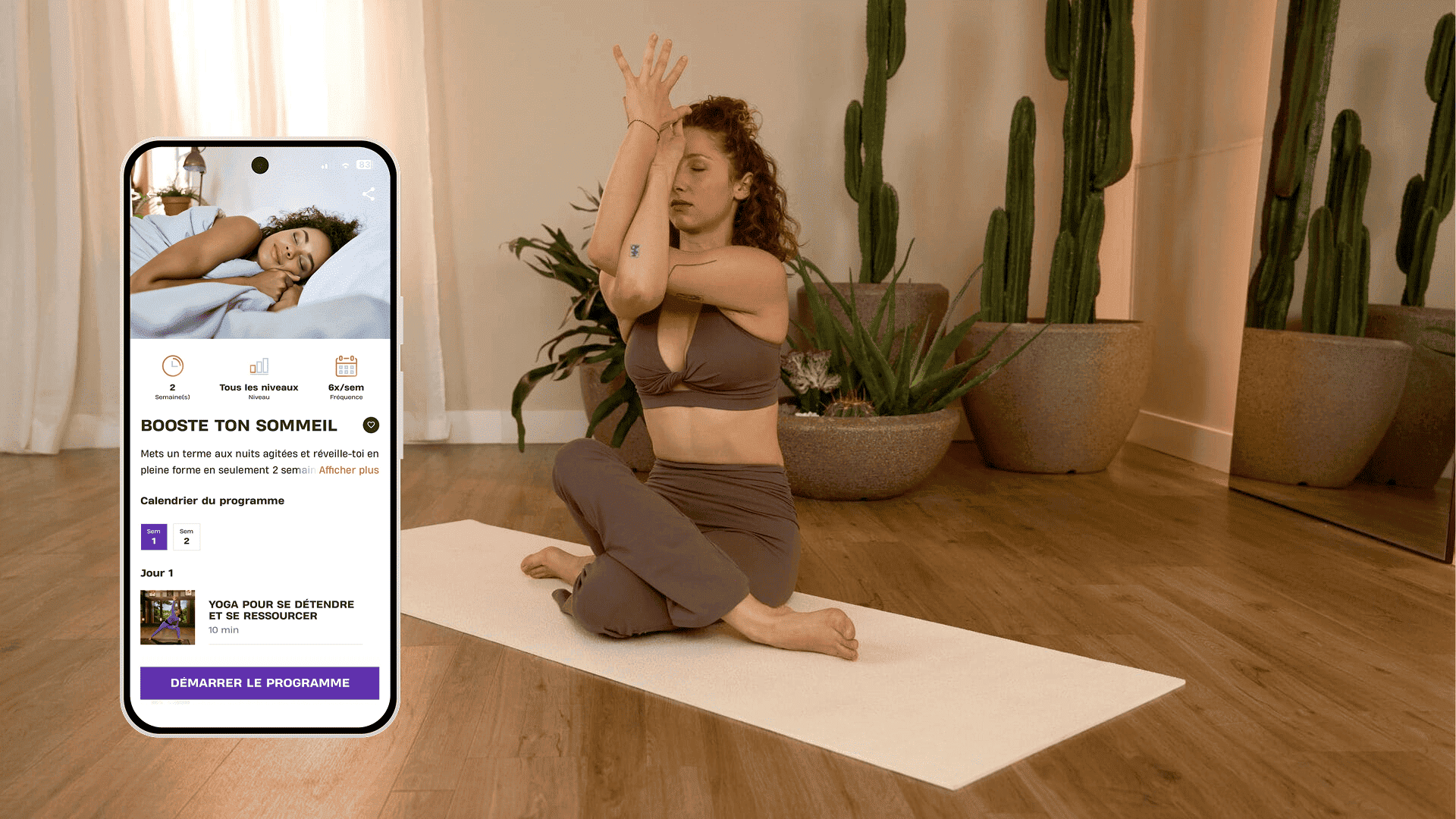 Une femme pratique une posture de yoga sur un tapis dans une pièce calme et lumineuse décorée de grandes plantes. Elle est assise en tailleur et se concentre sur un étirement des bras et des épaules. À côté d’elle, un smartphone affiche le programme Basic-Fit « Booste ton sommeil », présentant des séances de yoga pour se détendre et mieux dormir.