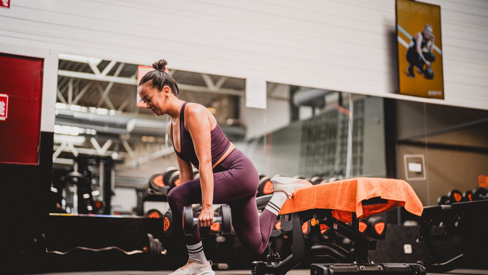 Femme en tenue de sport violette effectuant un squat bulgare avec haltères sur un banc dans la salle de sport Basic-Fit.