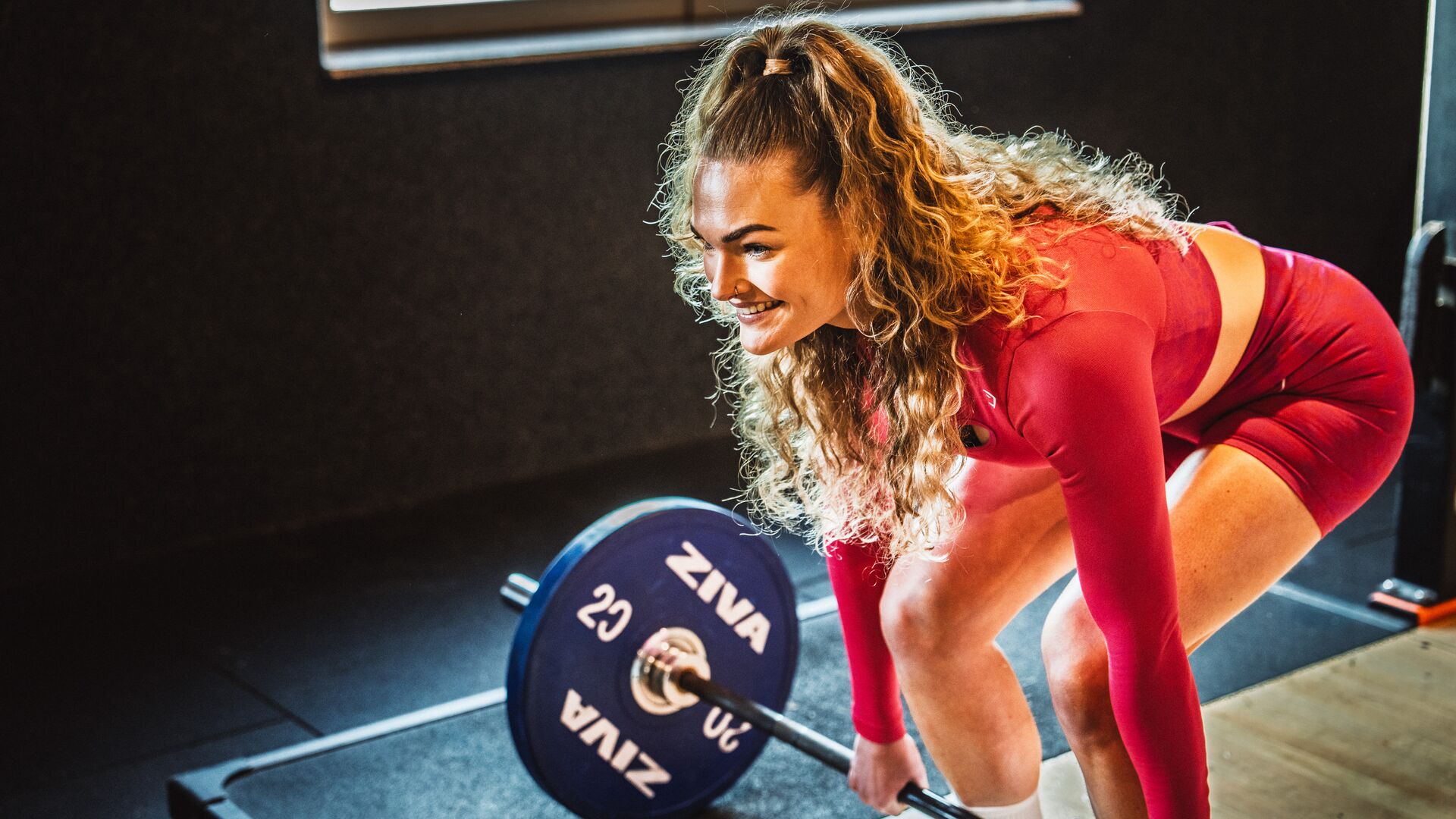 A woman in a red sports outfit is lifting a barbell in a Basic-Fit gym while smiling. Warm lighting highlights her from the side.