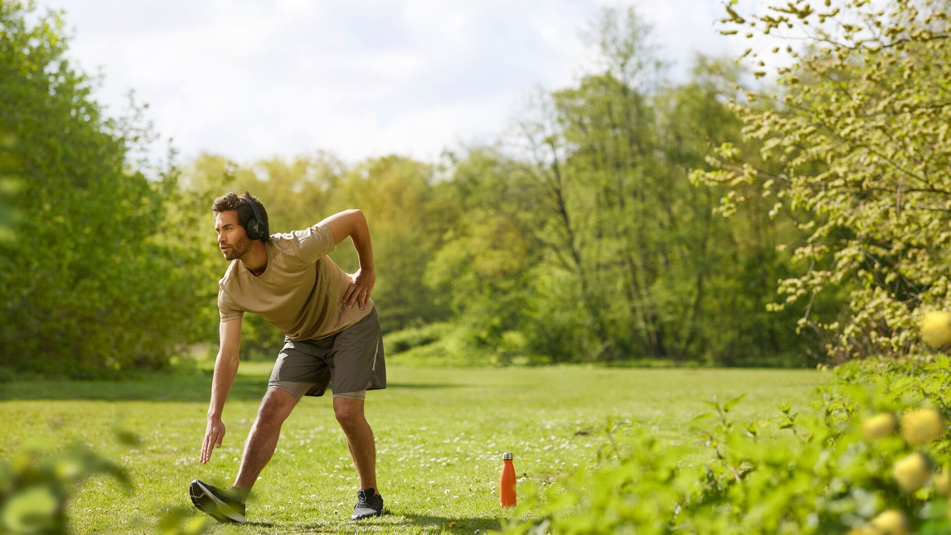 Un homme en t-shirt beige et short s’étire dehors sur l’herbe, portant un casque audio, avec une gourde orange posée à côté de lui.