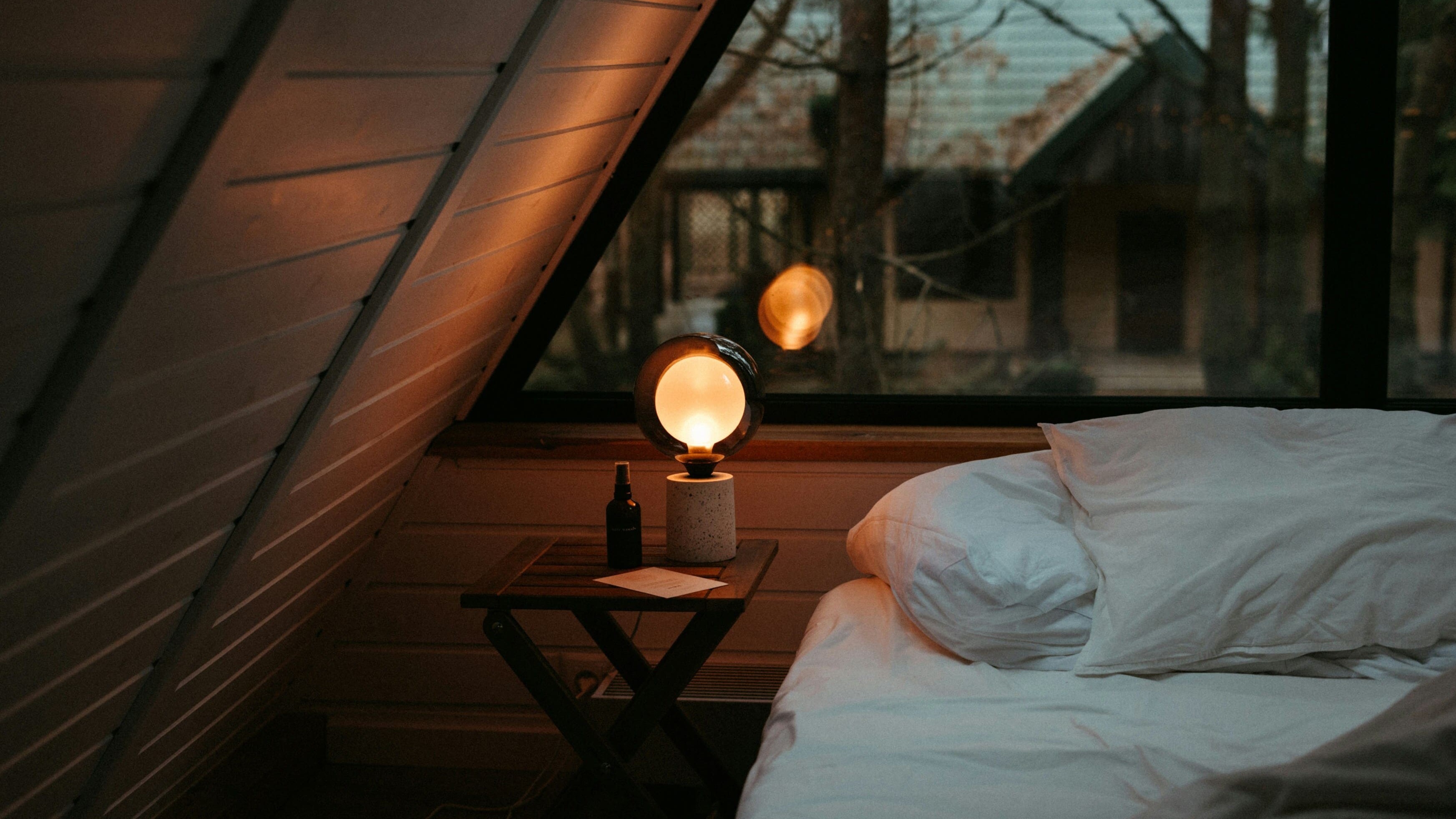 Une chambre mansardée chaleureuse avec du linge de lit blanc et une lampe en bois allumée, créant une ambiance calme et apaisante.