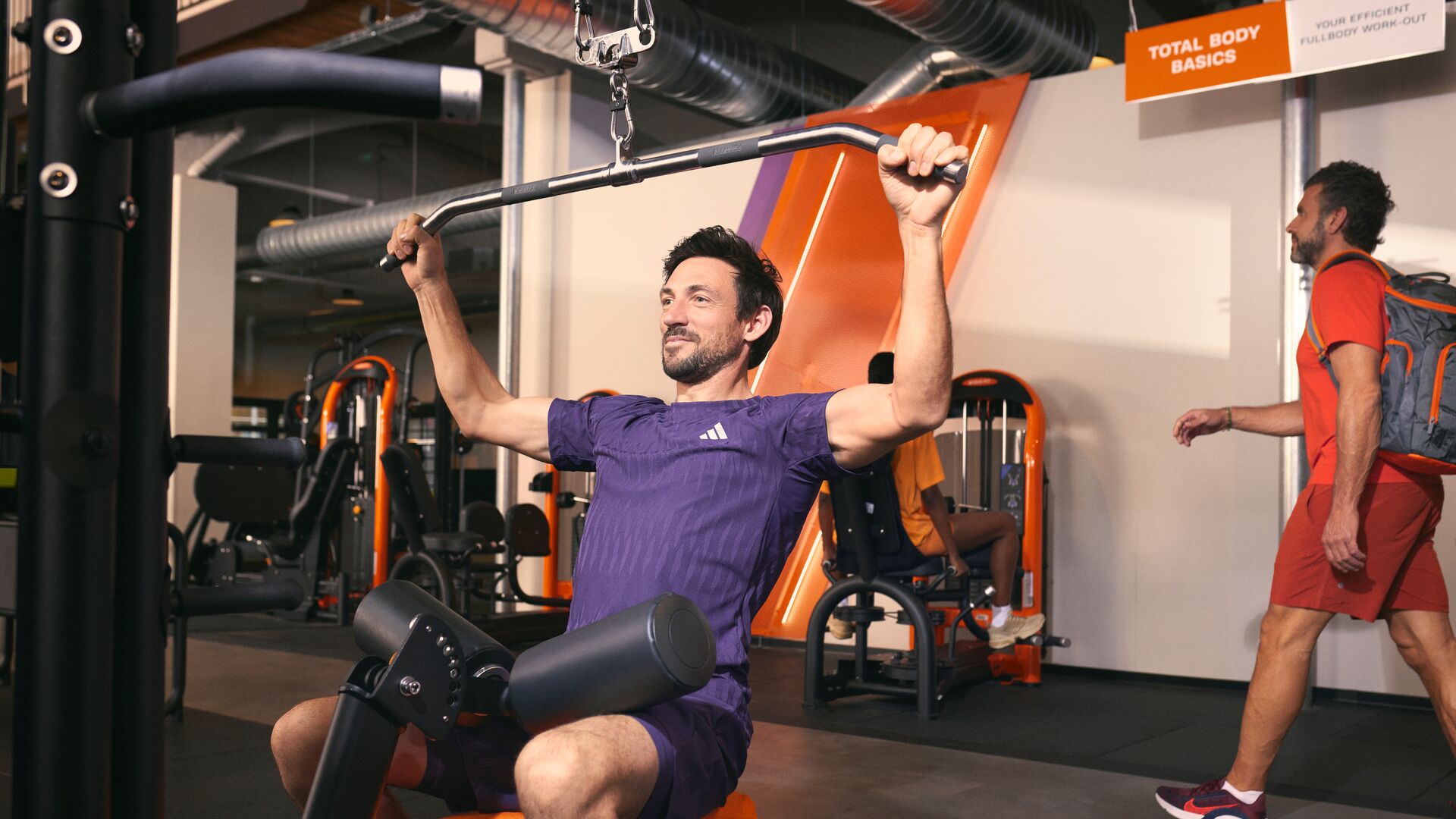 Homme réalisant un exercice de lat pulldown sur une machine dans une salle de sport Basic-Fit moderne.