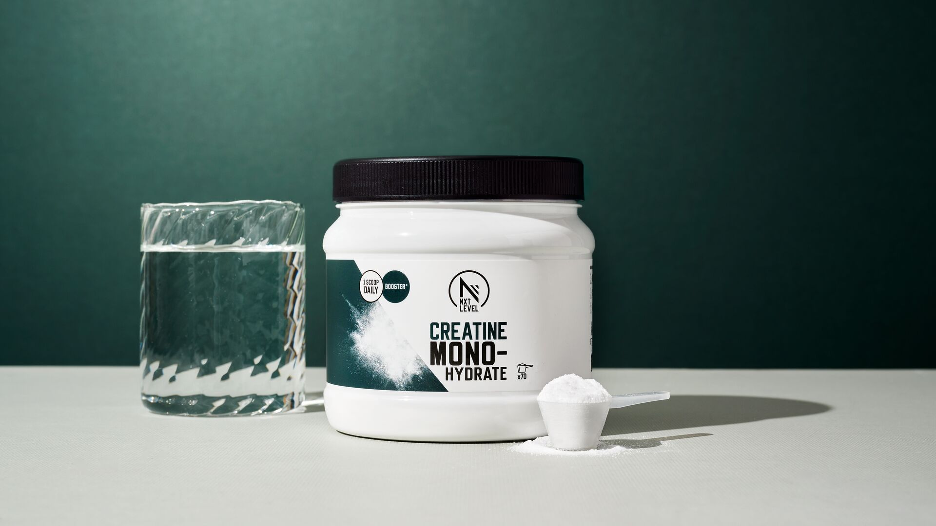 Un pot de créatine monohydrate est posé sur une table à côté d’un verre d’eau et d’une dosette remplie de poudre, photographié sur un fond vert foncé.