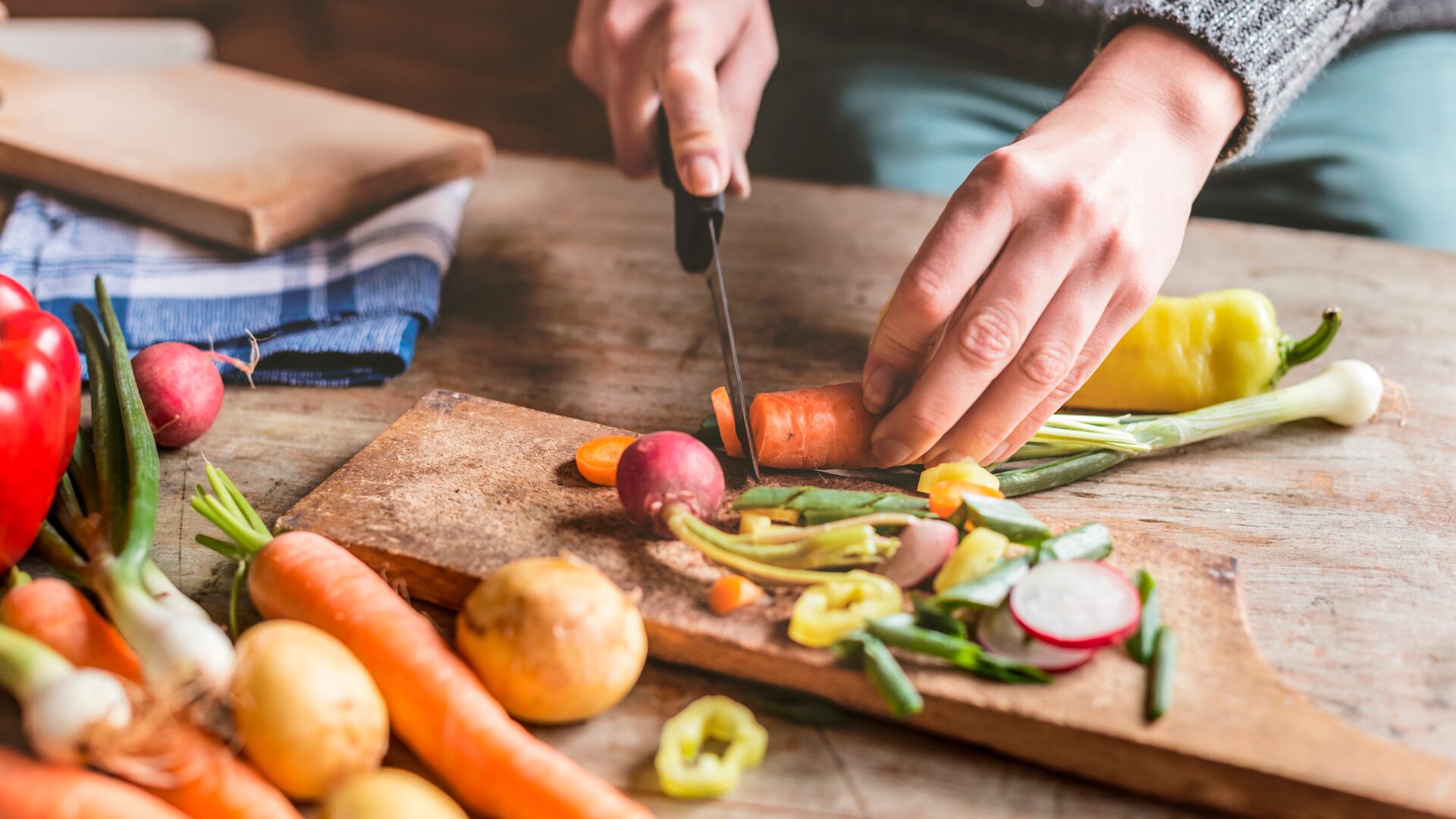 Gros plan sur des mains en train de couper des légumes frais sur une planche en bois, entourées d’ingrédients colorés comme des carottes, pommes de terre, radis et poivrons.