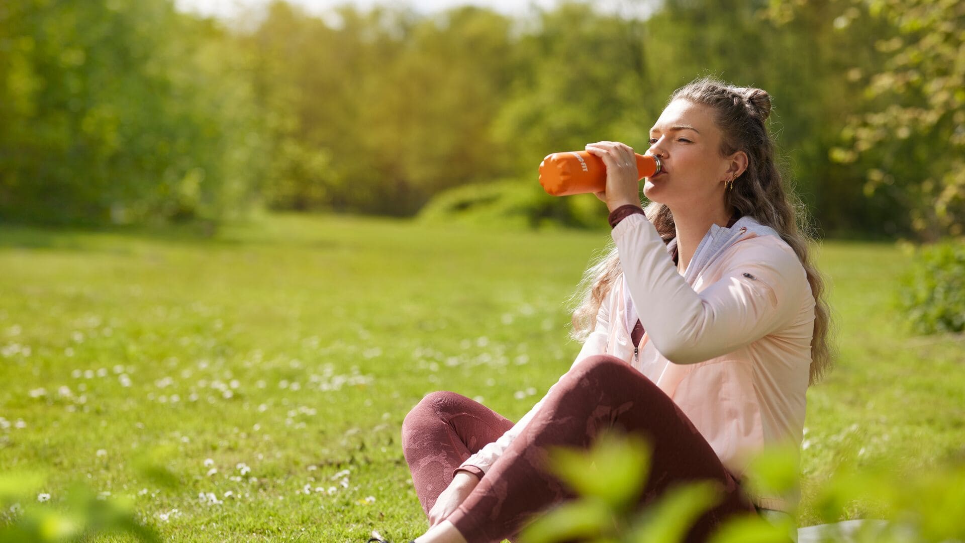 Une femme en veste légère et en legging bordeaux est assise dans l’herbe d’un parc ensoleillé et boit dans une gourde orange après son entraînement.