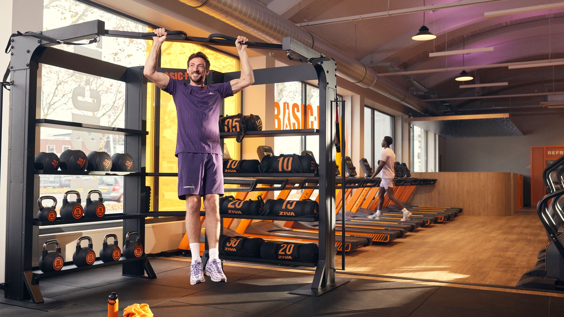 Personne effectuant une traction à la barre dans une salle de sport, à proximité des poids libres et des tapis de course, avec la lumière du jour entrant par de grandes fenêtres.