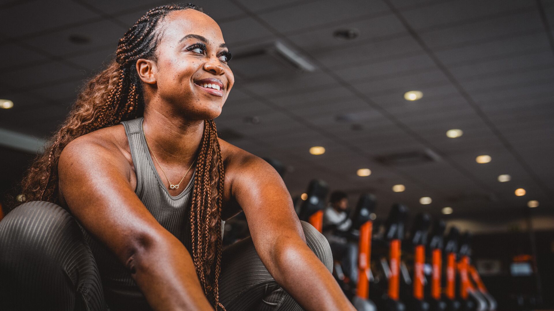 Vrouw in een bruine sportoutfit die op de vloer van de sportschool zit en glimlacht, met cardioapparaten en oranje accenten op de achtergrond.