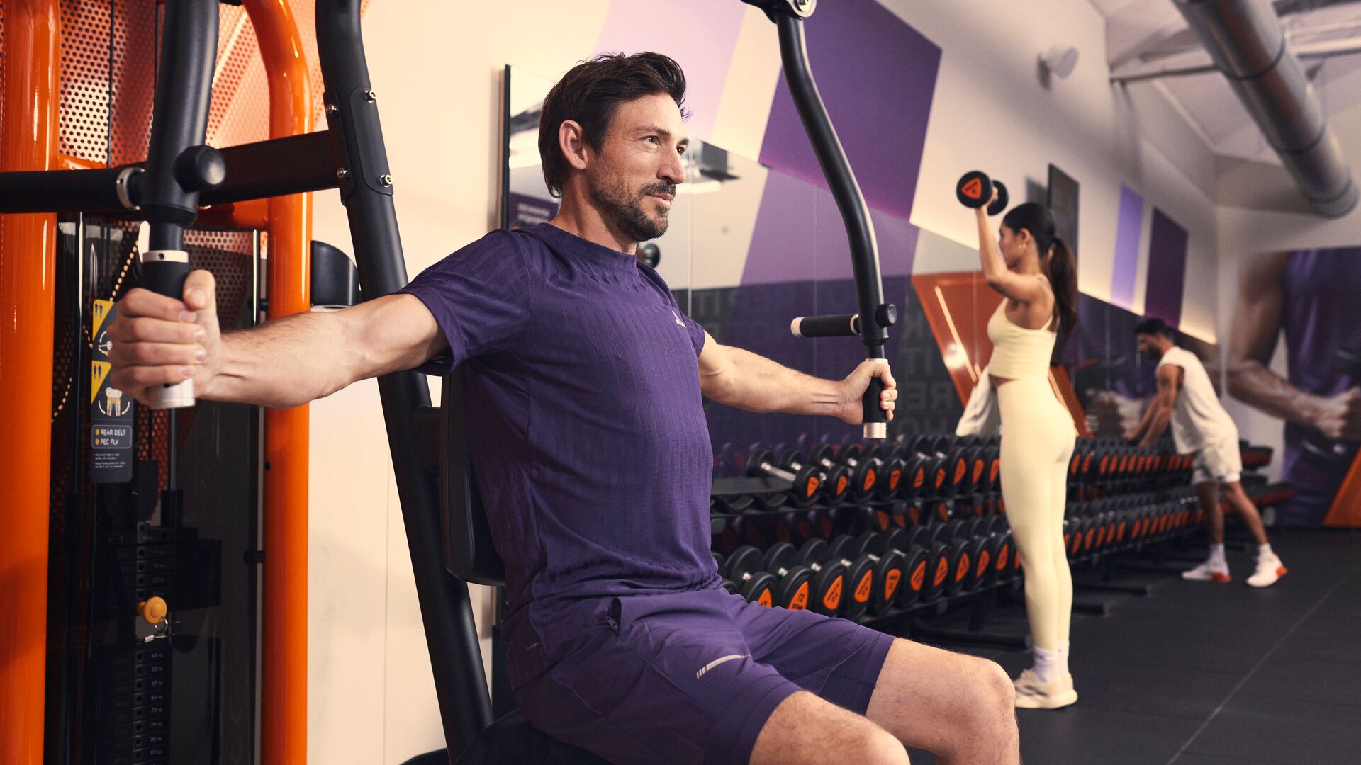Homme utilisant une machine de chest fly dans une salle de sport pendant que d’autres personnes s’entraînent en arrière-plan.