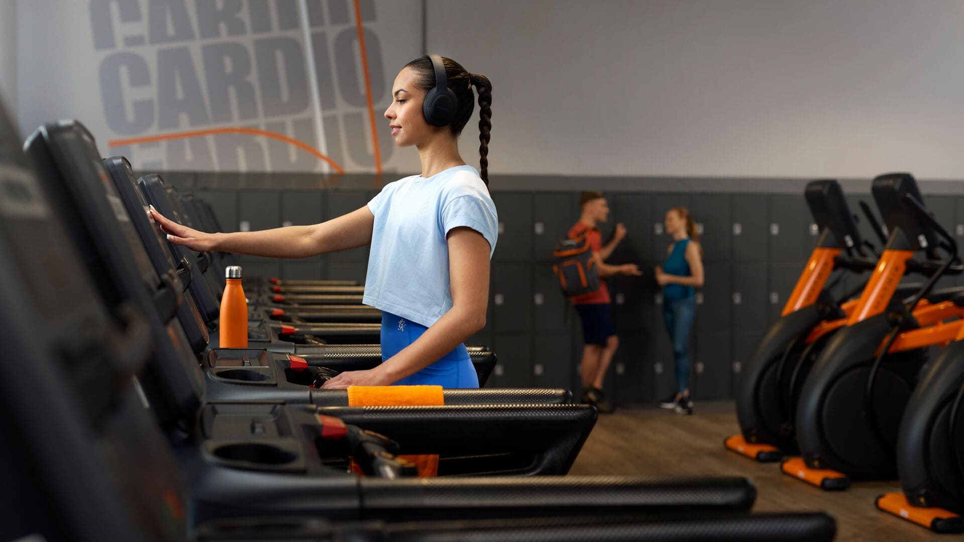 Une femme en haut bleu clair et legging bleu utilise un tapis de course chez Basic-Fit, portant un casque audio, avec une gourde orange posée devant elle.