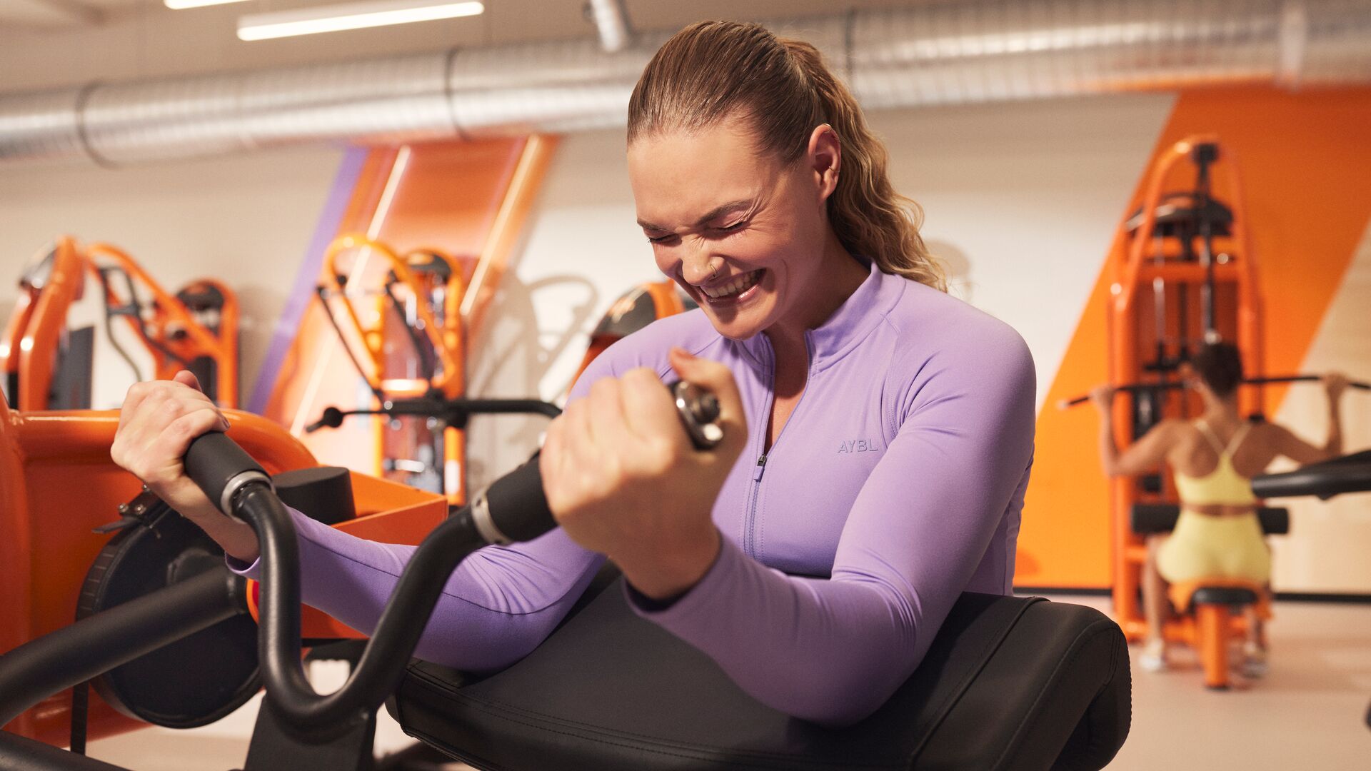 Une femme réalise un exercice de musculation assise sur une machine dans une salle Basic-Fit, concentrée sur son entraînement tout en souriant. Des équipements de musculation et des touches orange sont visibles en arrière-plan.