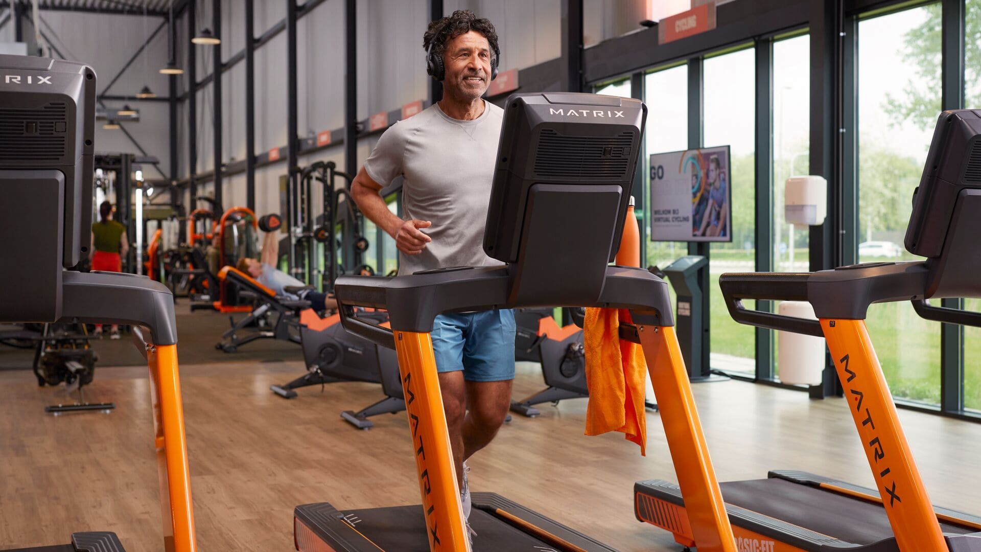 A man in a gray T-shirt and blue shorts is jogging on a treadmill in a Basic-Fit gym, smiling and wearing headphones. Other members are visible in the background.