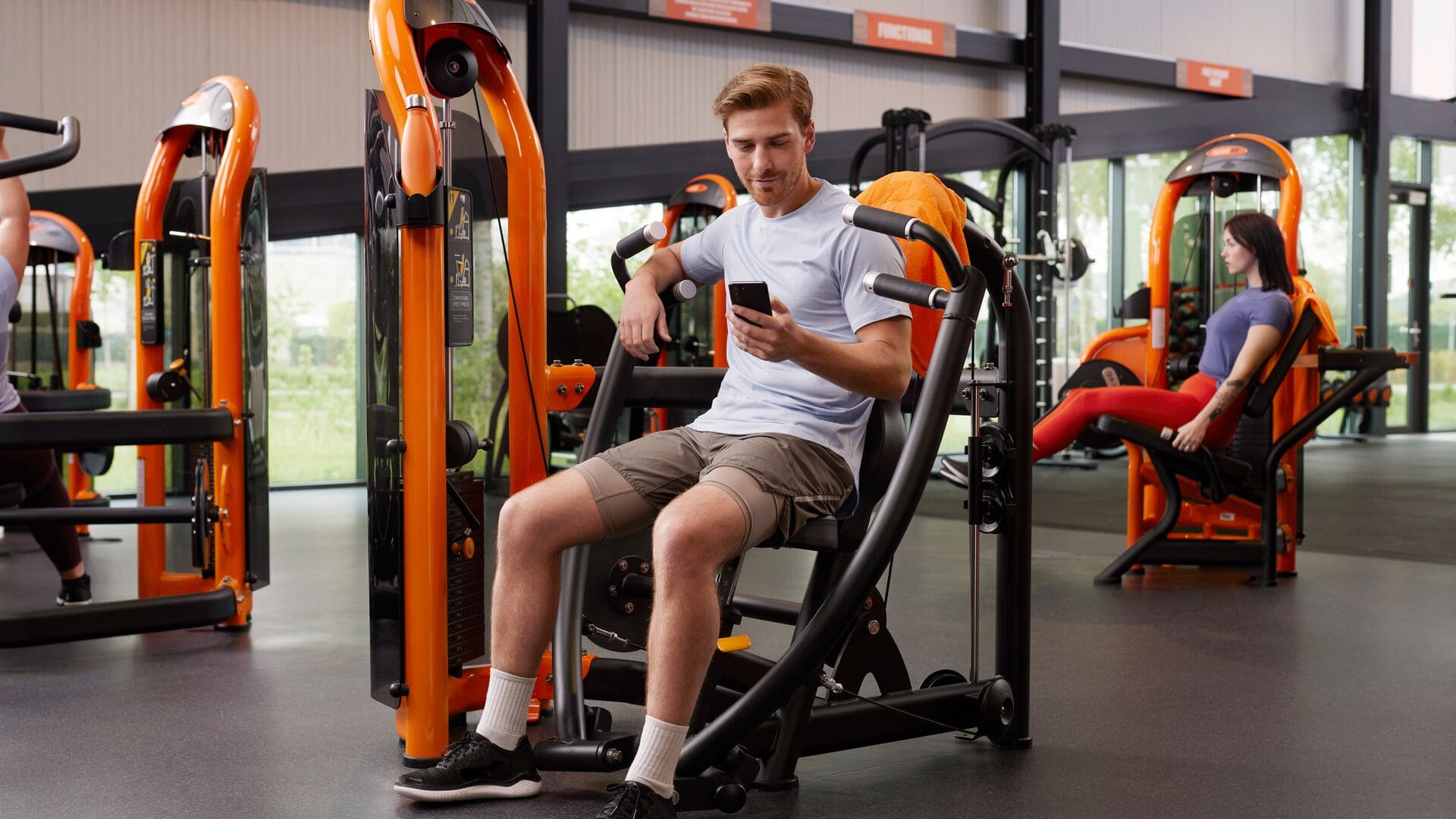 Homme assis sur une machine de développé couché regardant son téléphone dans la salle de sport Basic-Fit, avec d’autres membres utilisant des machines de musculation en arrière-plan.