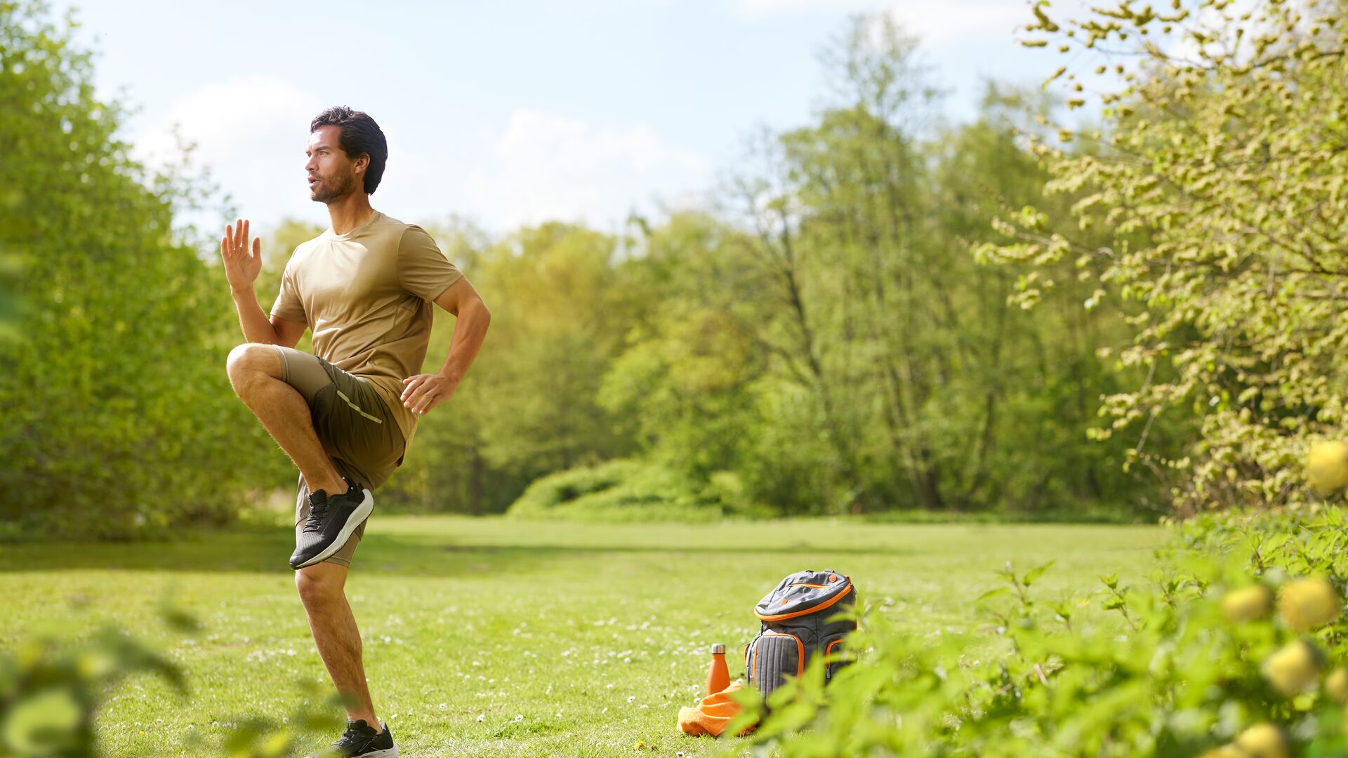 Un homme fait un exercice de genoux hauts dans un parc ensoleillé, avec un sac à dos, une bouteille d’eau et une serviette posés sur l’herbe à côté.