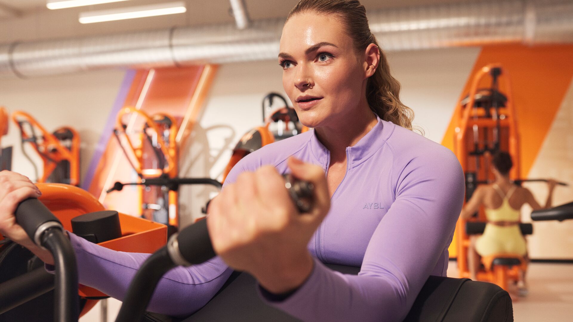 Personne utilisant une machine de curl biceps en position assise dans une salle de sport, concentrée sur le mouvement des bras, avec des machines orange en arrière-plan.