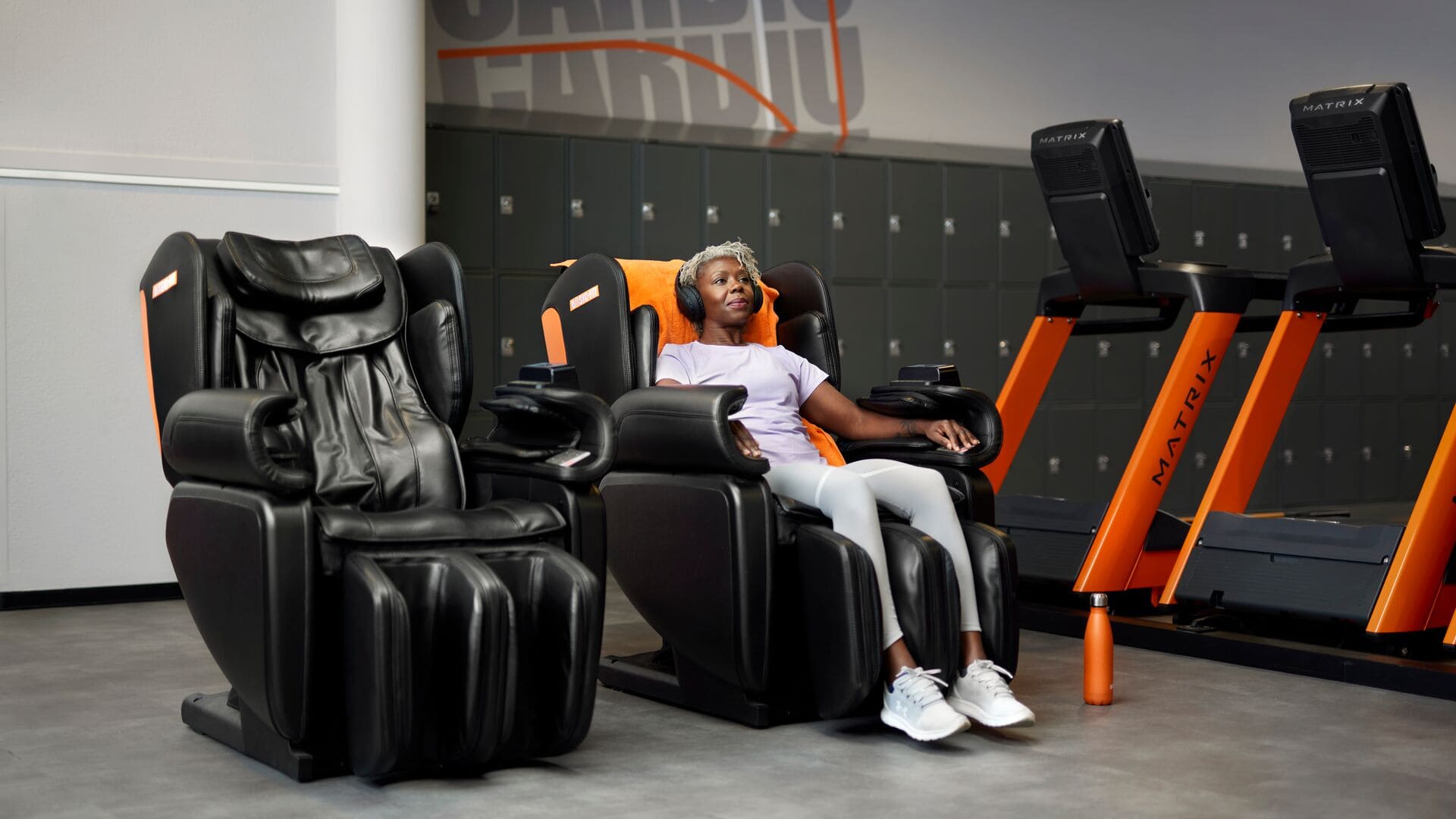 Une femme se détend dans un fauteuil de massage noir avec des accents orange, casque audio sur les oreilles, dans un club Basic-Fit. Elle est assise confortablement après son entraînement, avec des tapis de course et des casiers en arrière-plan.
