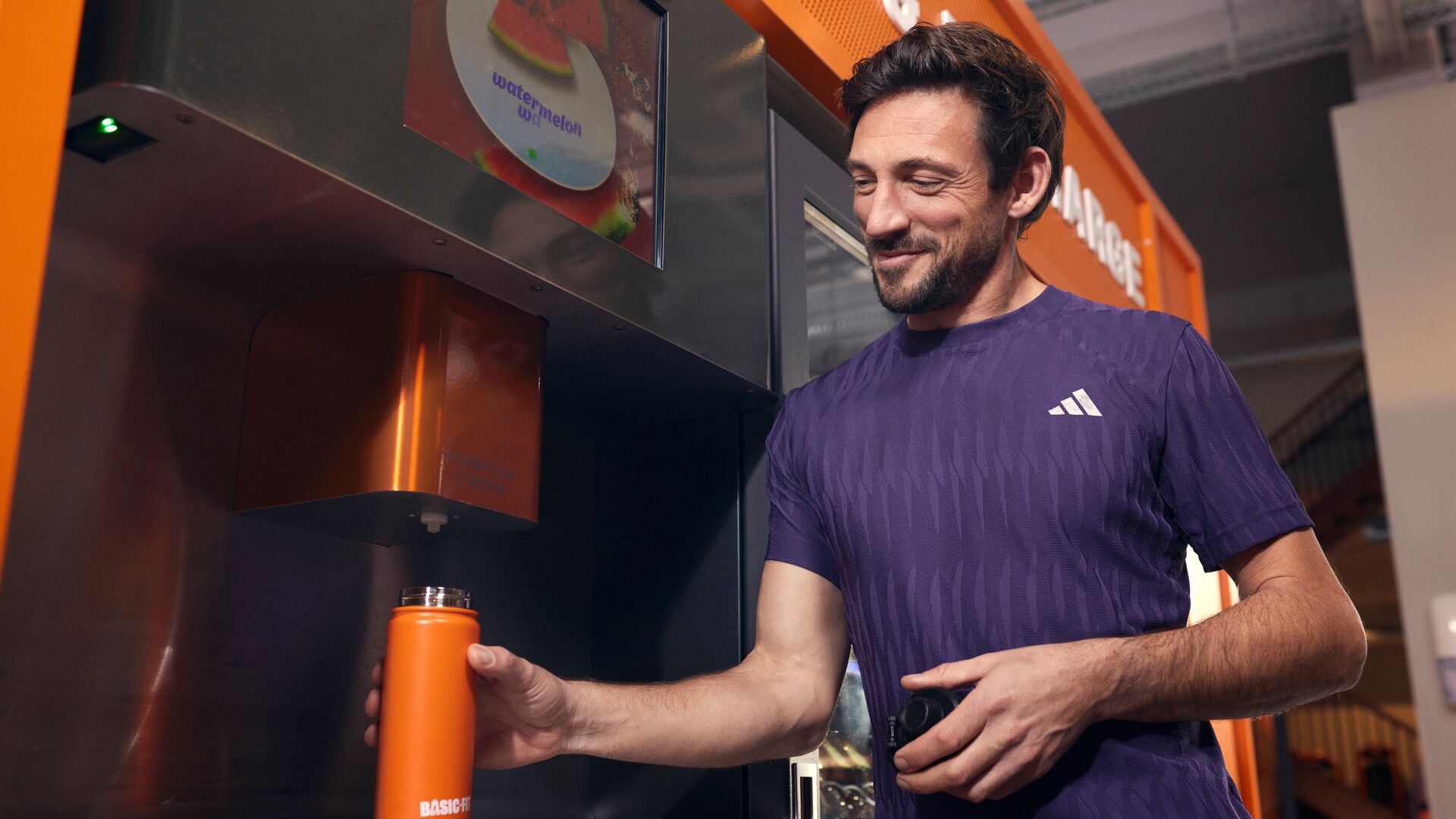 Un homme remplit une gourde orange Basic-Fit à une borne de boissons en libre-service dans une salle Basic-Fit.
