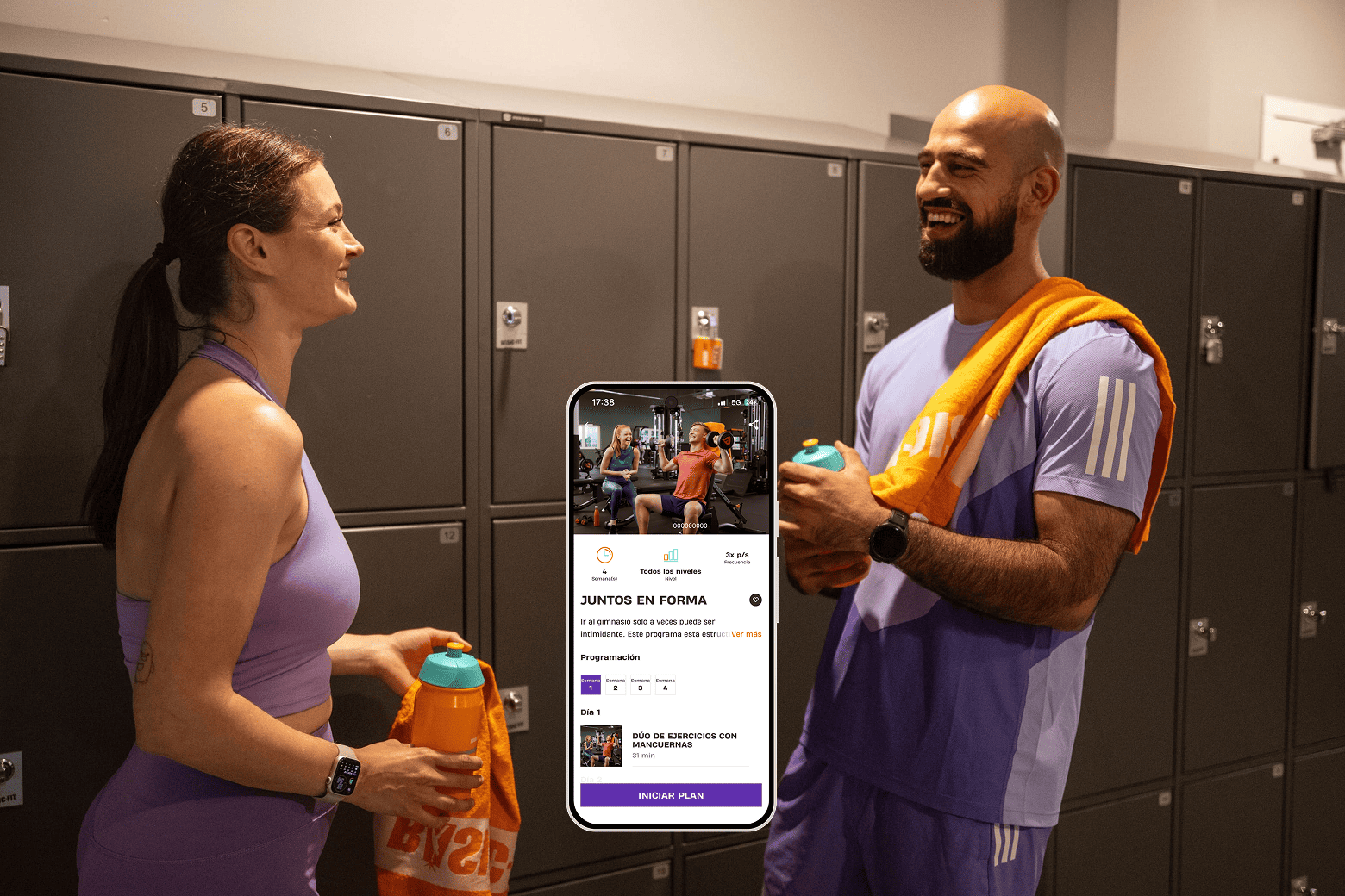 Dos personas en el vestuario del gimnasio sonriendo mientras sostienen botellas de agua. En el centro hay un teléfono móvil que muestra una aplicación de entrenamiento llamada “Juntos en Forma” con un programa de ejercicios en pareja.