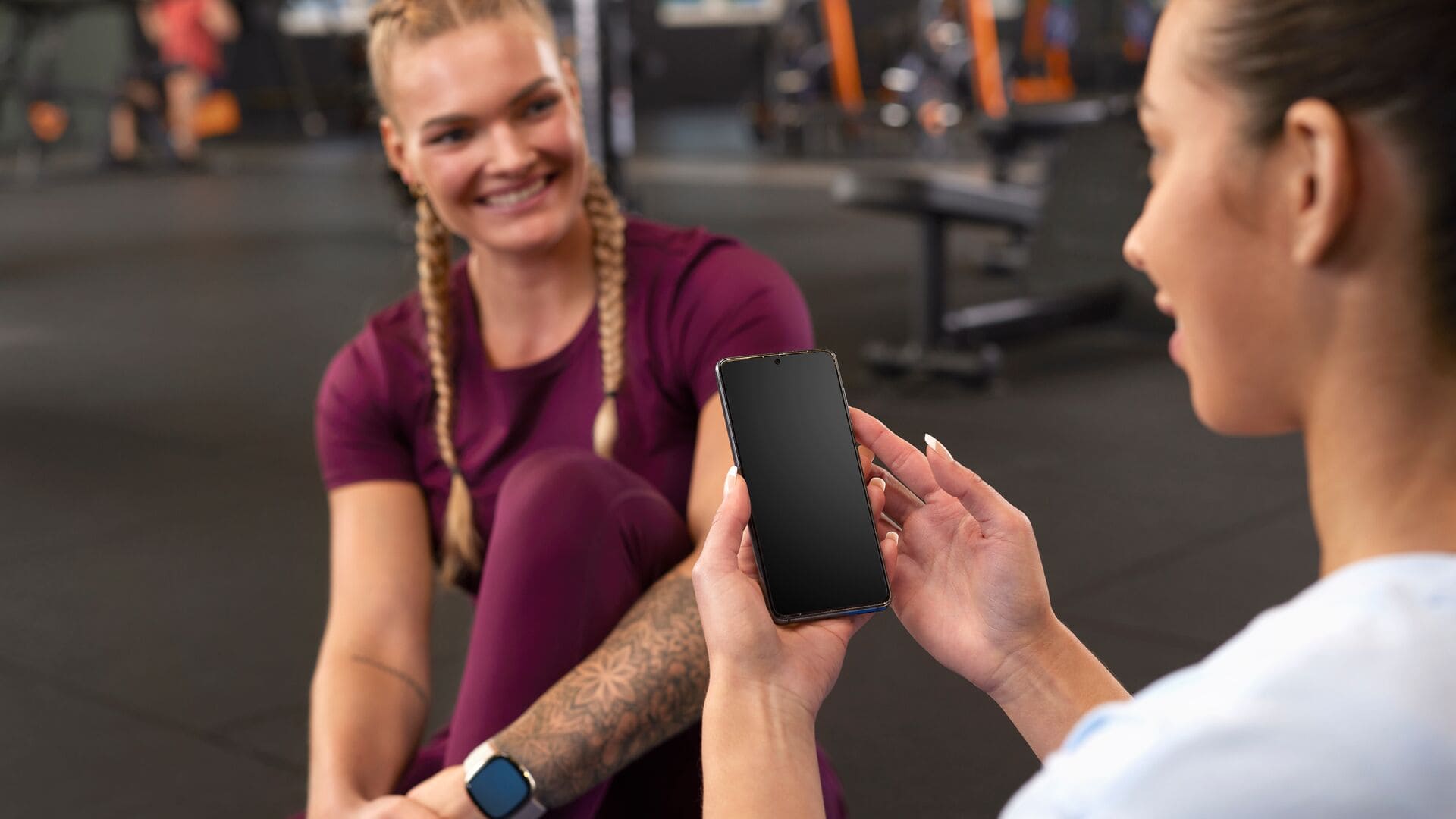 Deux femmes assises au sol de la salle de sport après l’entraînement ; l’une tient un smartphone en l’air tandis que l’autre sourit, toutes deux en tenue de sport.