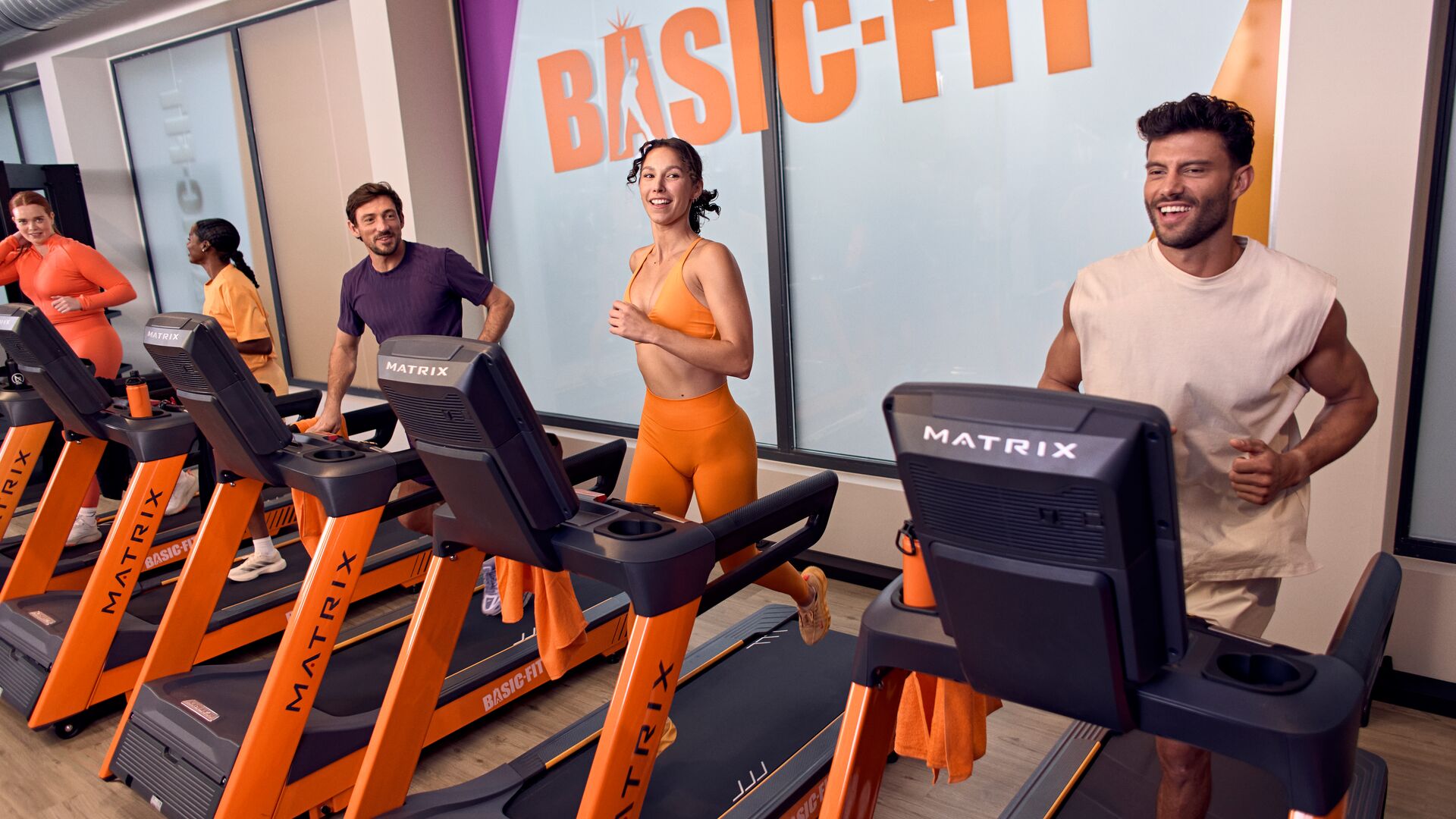 Groupe de personnes courant ensemble sur des tapis de course dans une salle Basic-Fit, dans un espace cardio lumineux.