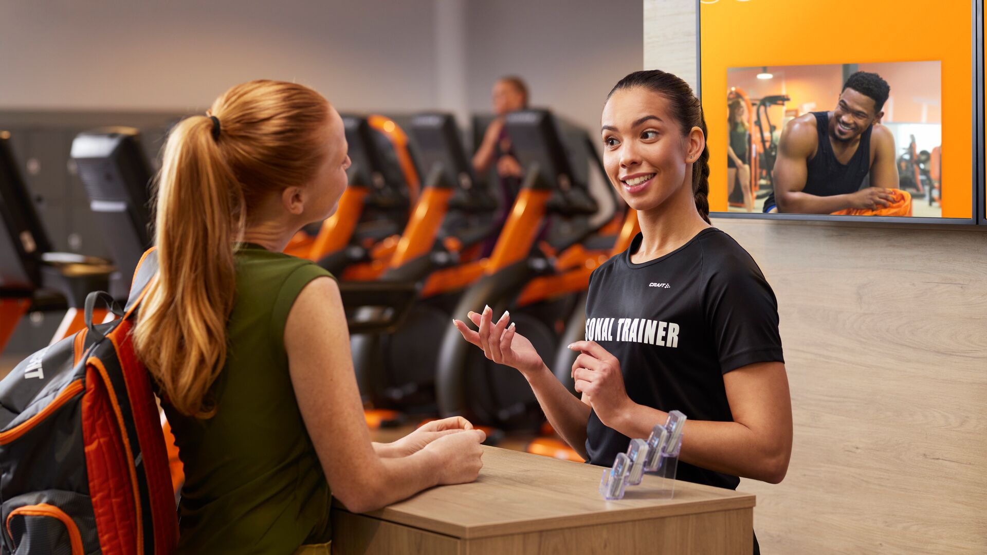 Un coach personnel dans une salle Basic-Fit accueille chaleureusement un membre à l’accueil, avec des machines de cardio orange et un écran numérique en arrière-plan.