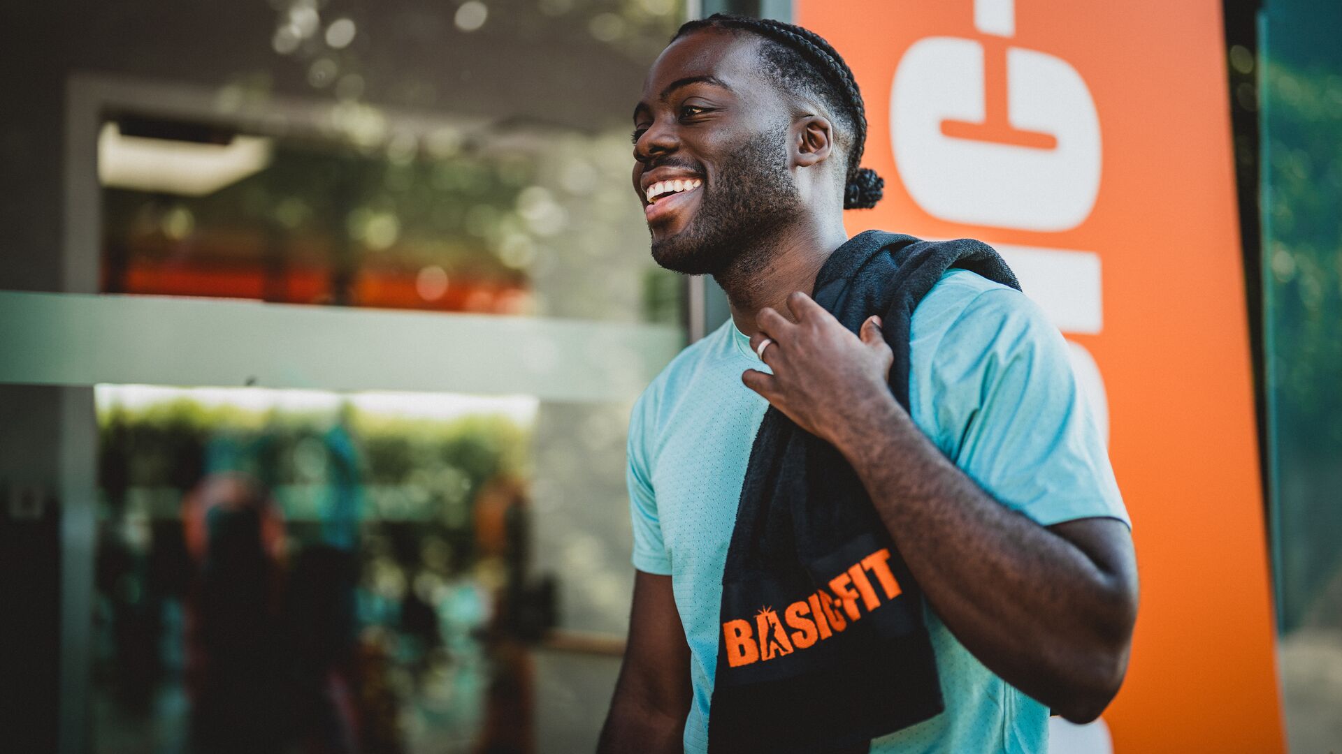 Lachende man in een lichtblauw T-shirt buiten bij een Basic-Fit club, met een zwarte handdoek met oranje Basic-Fit logo over zijn schouder.