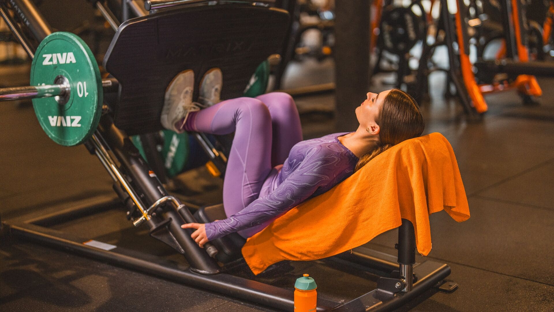 Une femme en tenue de sport violette s’entraîne sur une machine de leg press dans une salle Basic-Fit, avec une serviette orange posée sur le banc et des poids à proximité.