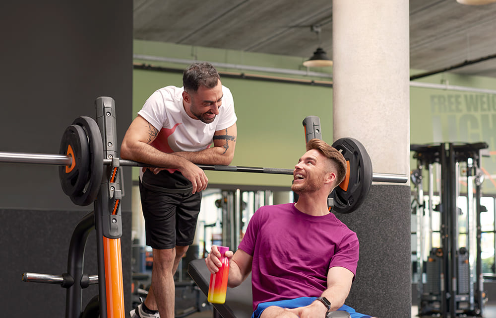 Two men are in a Basic-Fit gym talking and laughing near a barbell on a weight rack. One is standing in a white and red shirt, and the other is sitting in a purple shirt holding a shaker bottle.