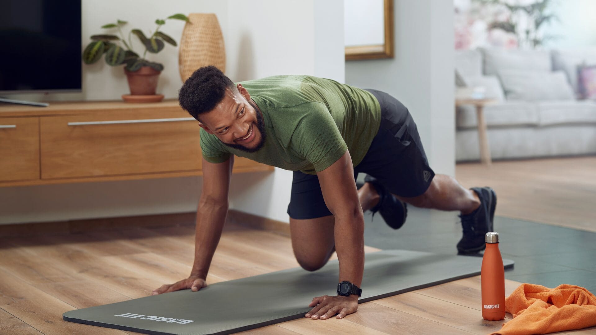 A man wearing a green T-shirt and black shorts is exercising on a gray Basic-Fit mat at home, smiling while holding a plank position. An orange Basic-Fit water bottle and a towel are on the floor beside him.