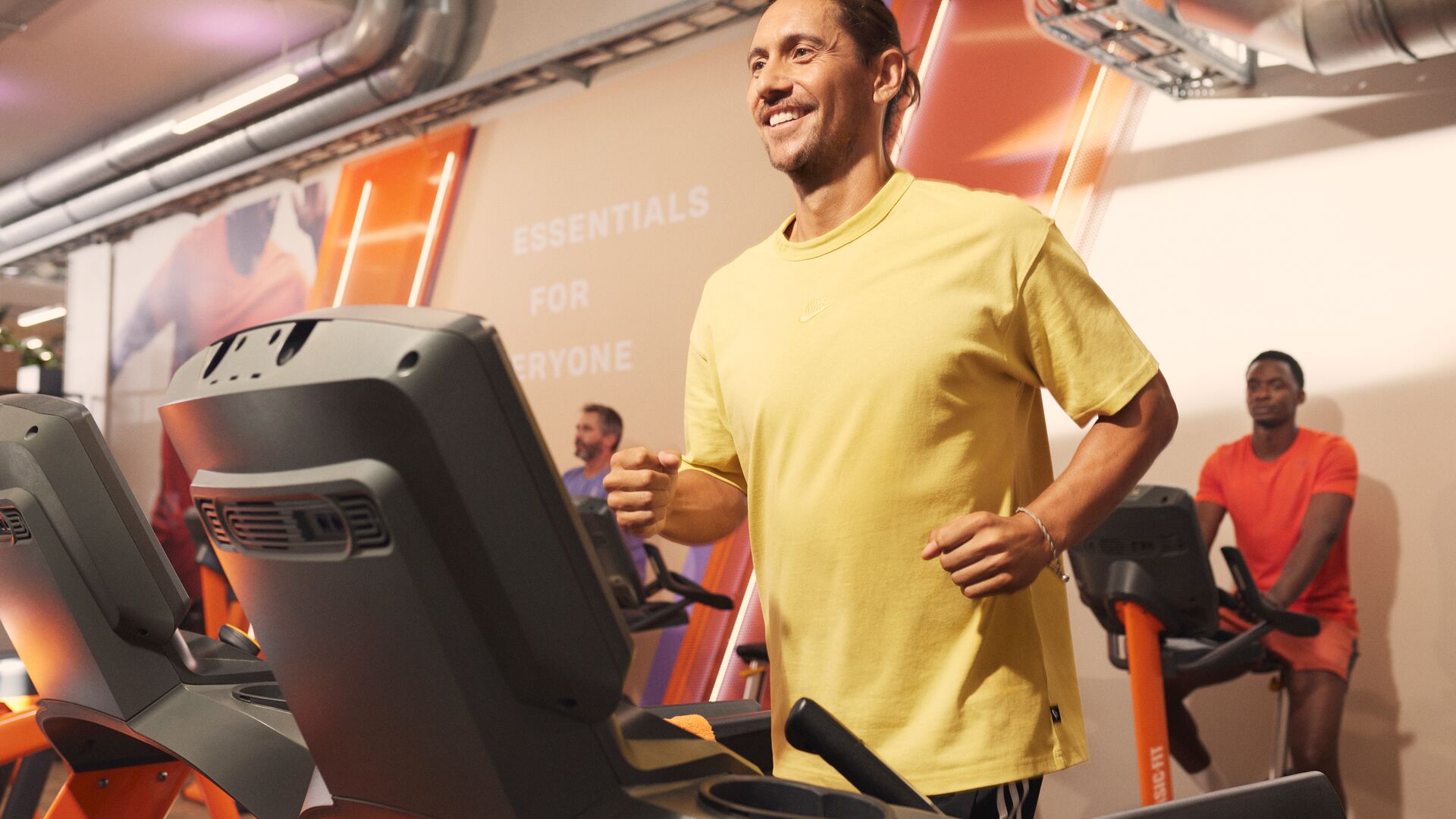 Un homme court sur un tapis de course dans une salle Basic-Fit, vêtu d’un t-shirt jaune, tandis que d’autres adhérents s’entraînent sur des machines de cardio derrière lui dans un espace lumineux avec des détails orange.