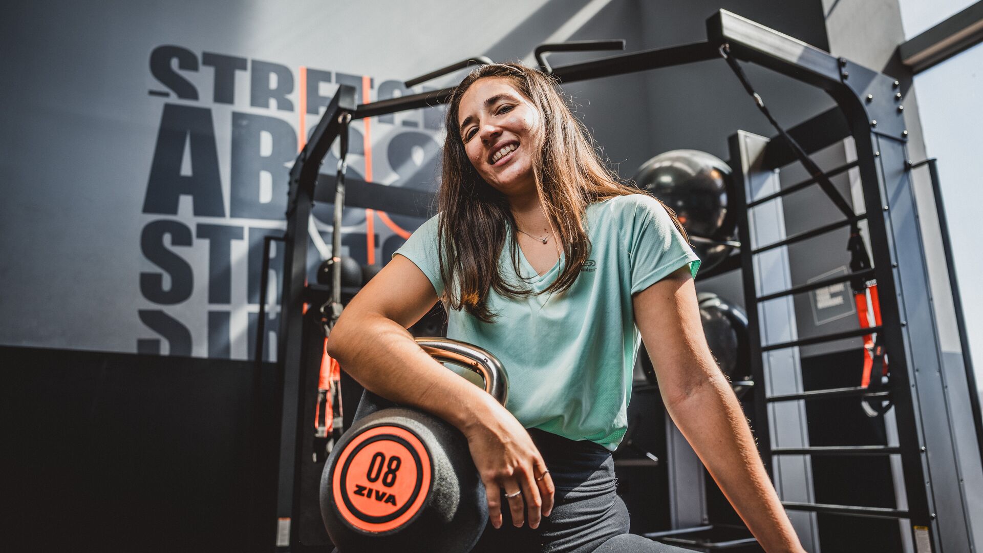Vrouw in een mintgroen T-shirt die in de sportschool zit, leunend op een kettlebell van 8 kg, glimlachend naar de camera.