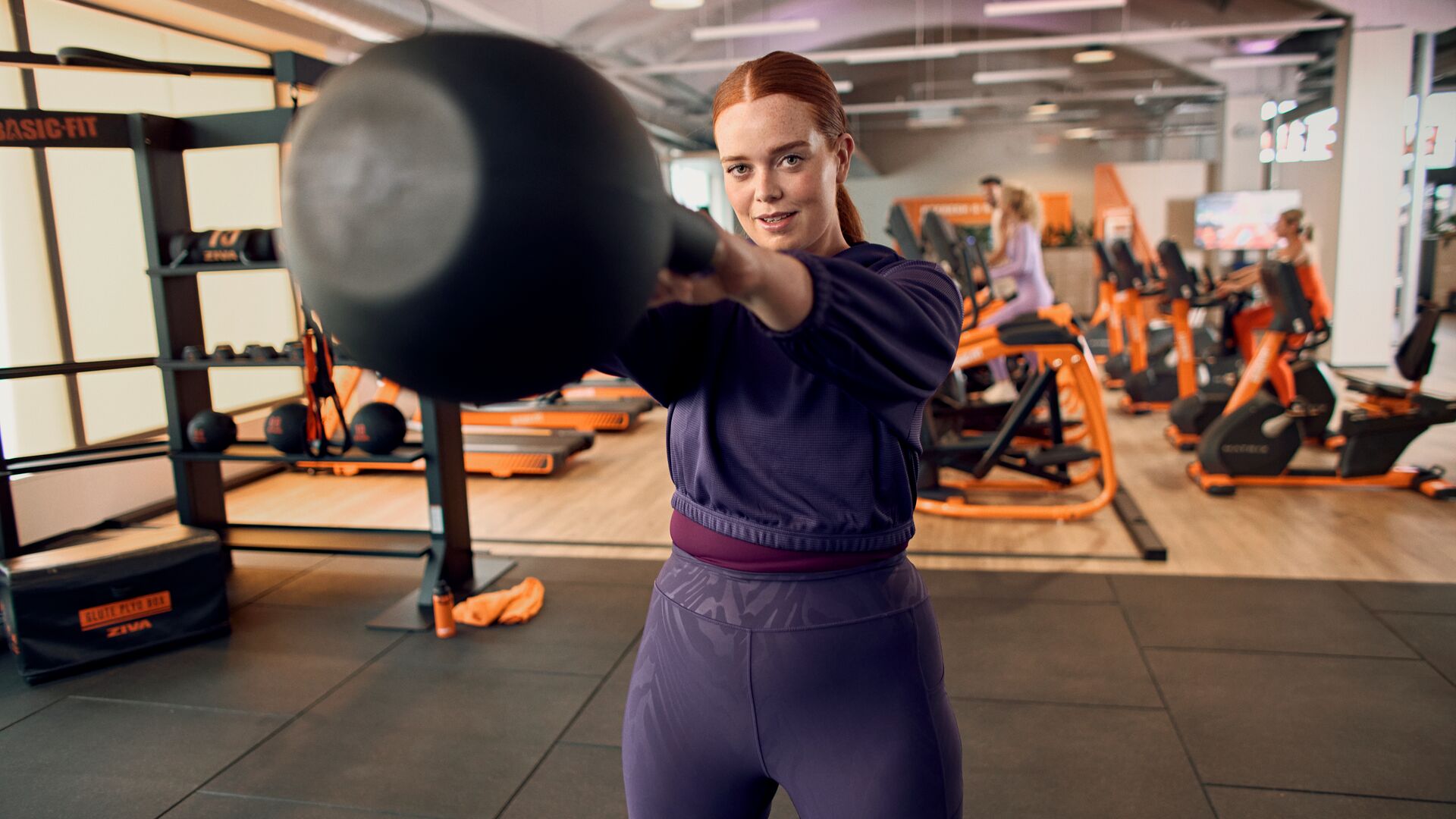 Femme s’entraînant avec un kettlebell dans une salle Basic-Fit, tenant le poids à hauteur d’épaule pendant un exercice de renforcement.