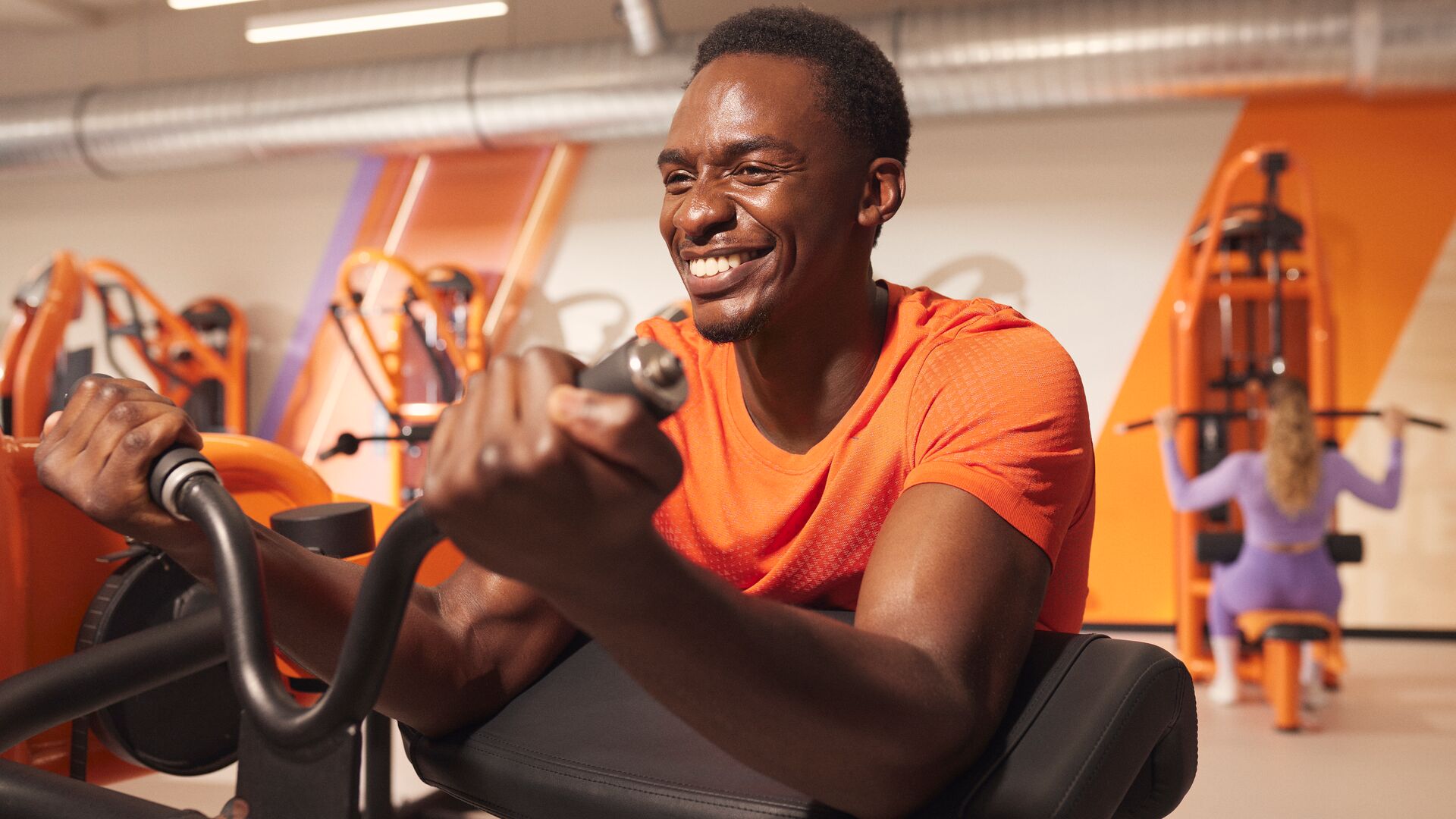 Personne réalisant un curl biceps assis sur une machine dans une salle de sport lumineuse, souriante pendant l’exercice, avec d’autres sportifs en arrière-plan.