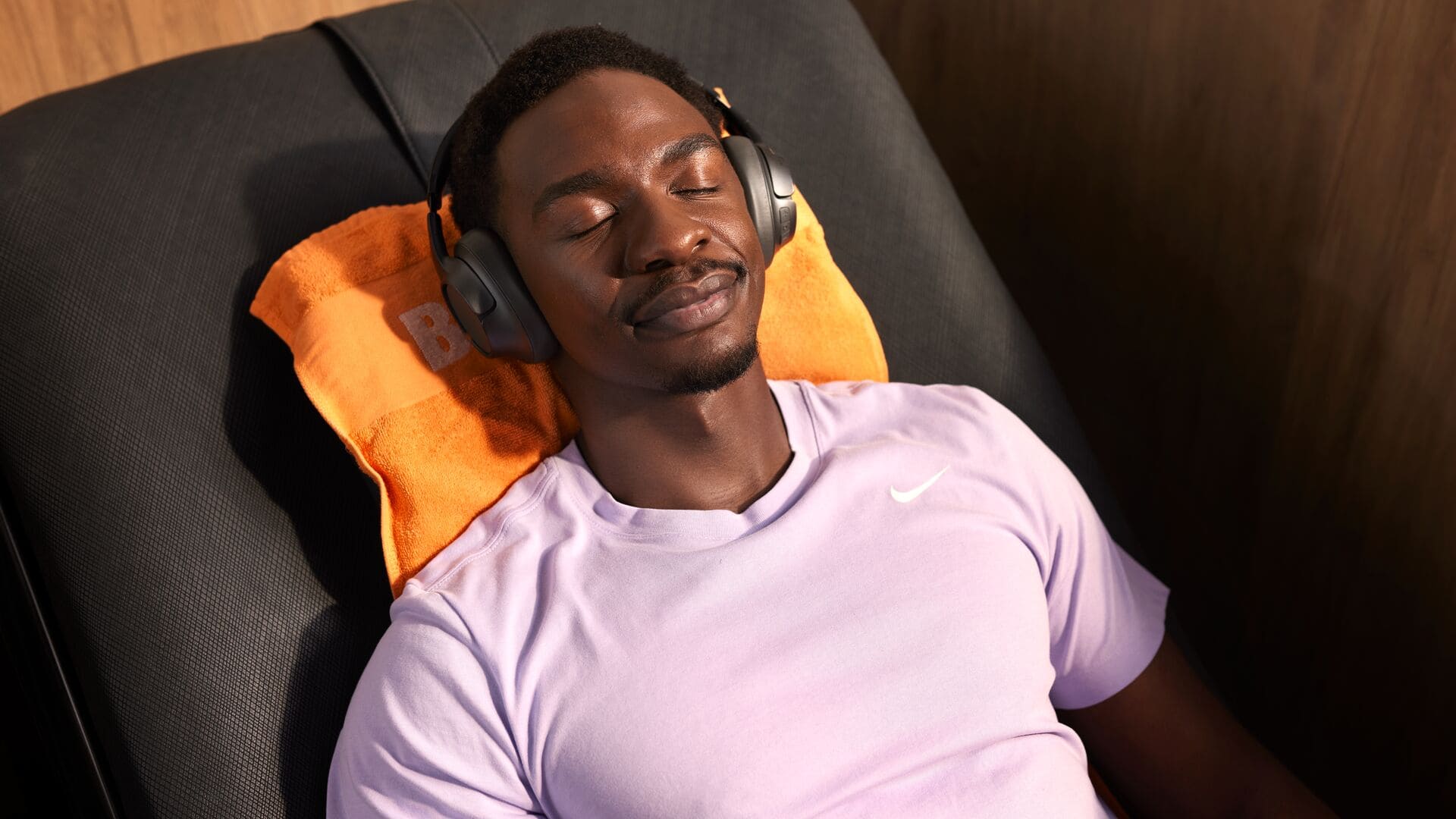Un homme en t-shirt de sport violet clair se détend avec un casque audio, allongé les yeux fermés sur une serviette orange dans un fauteuil de massage Basic-Fit.