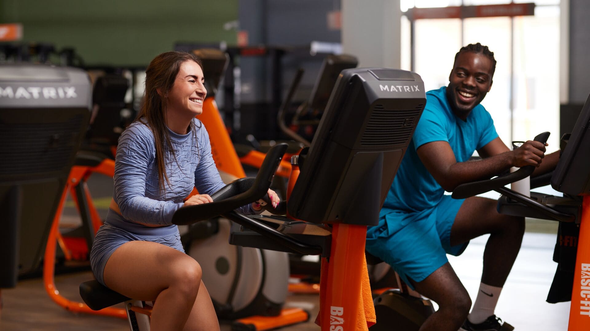 A woman in a light blue workout set and a man in a turquoise outfit are sitting on exercise bikes in a Basic-Fit gym, smiling and talking during their cardio session.