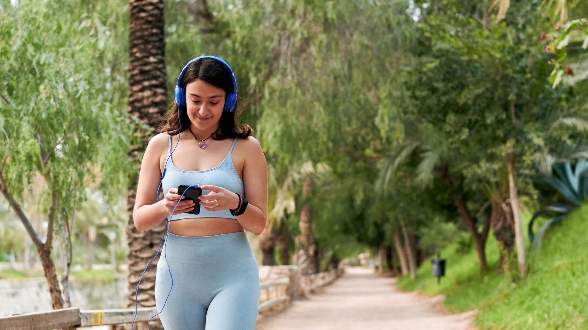 Une jeune femme en tenue de sport bleu clair marche à l’extérieur sur un chemin bordé d’arbres, avec un casque audio sur les oreilles et son téléphone à la main. Elle a l’air détendue et concentrée pendant sa marche ou sa séance.