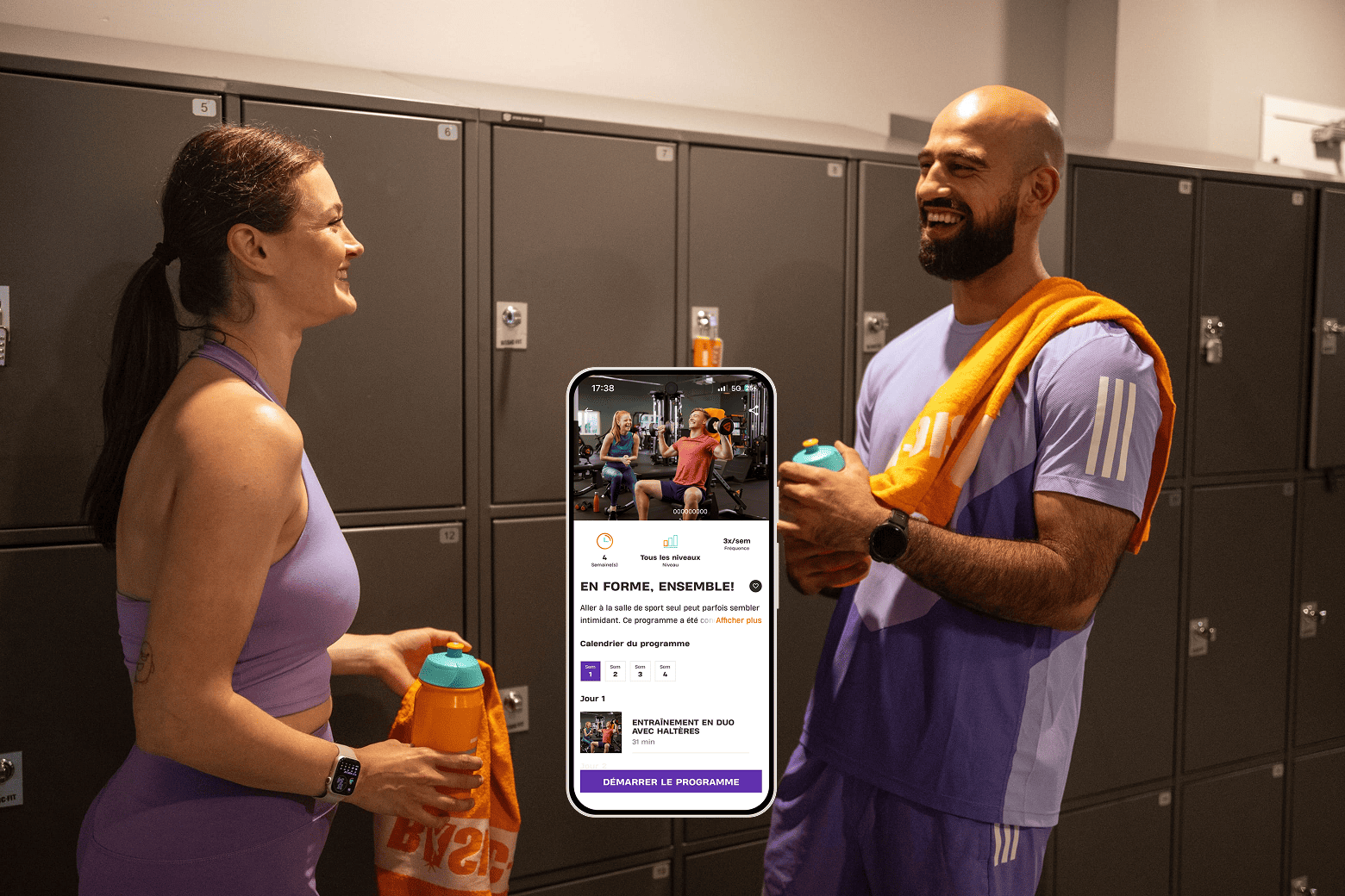 Deux personnes dans un vestiaire de salle de sport, souriantes et tenant des gourdes, l’une avec une serviette orange sur l’épaule. Entre elles, un écran de smartphone montre un programme d’entraînement en duo avec haltères intitulé “EN FORME, ENSEMBLE !” et un bouton violet “DÉMARRER LE PROGRAMME”.