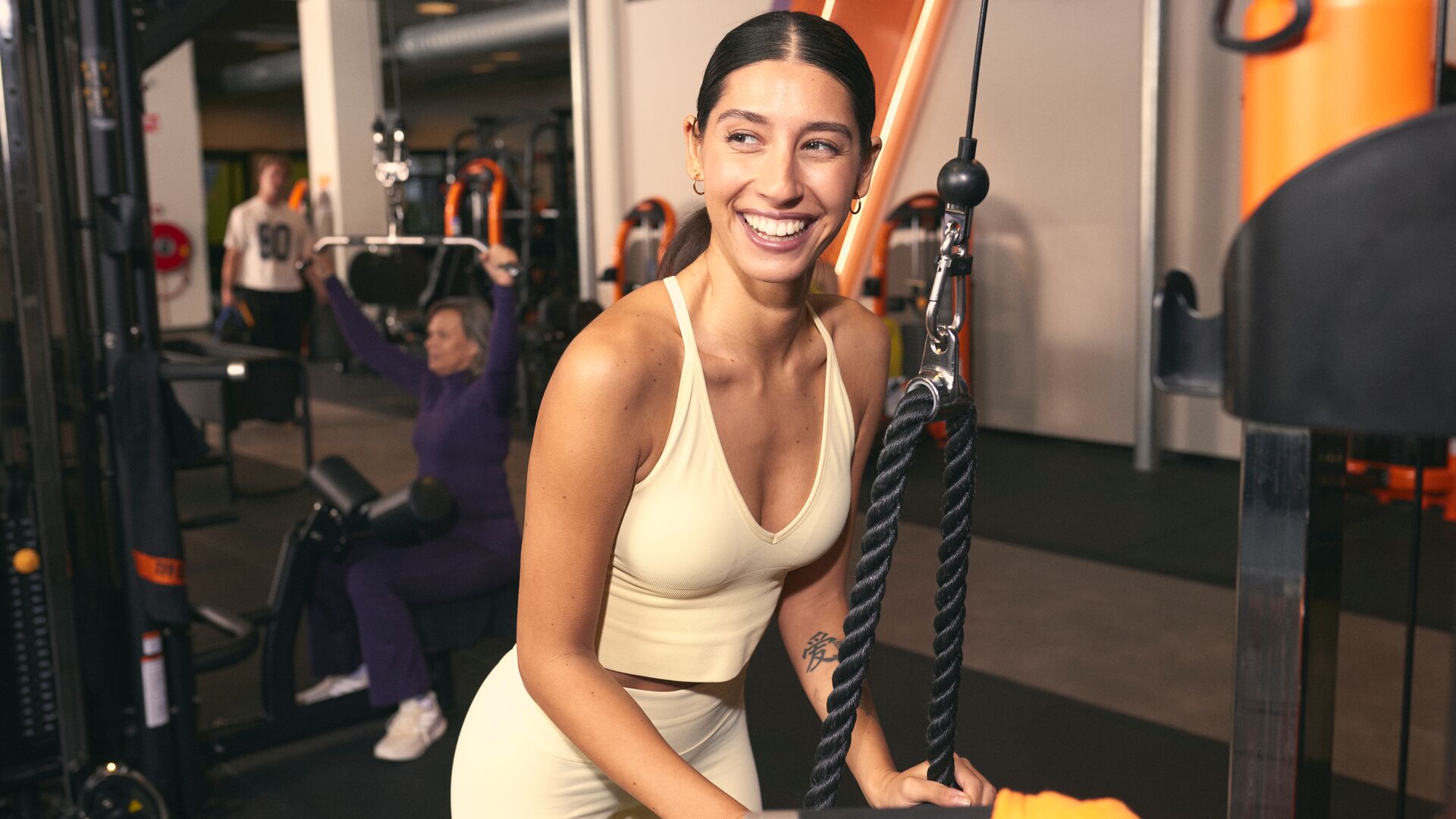 Une femme sourit en utilisant une machine à câbles dans une salle Basic-Fit, tenant une poignée corde pendant un exercice de musculation. D’autres adhérents s’entraînent en arrière-plan.